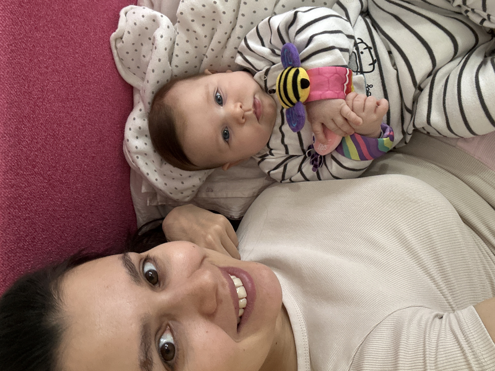 A woman and a baby are on a bed. The woman is in the foreground, smiling, and the baby is lying near her. The baby has blue eyes, light skin, and is wearing a black and white striped outfit. The baby is holding a bee-shaped toy. A white blanket with grey polka dots is near the baby. The bed has a pink section visible on the left side. The woman is wearing a light-colored shirt. The location is unknown.