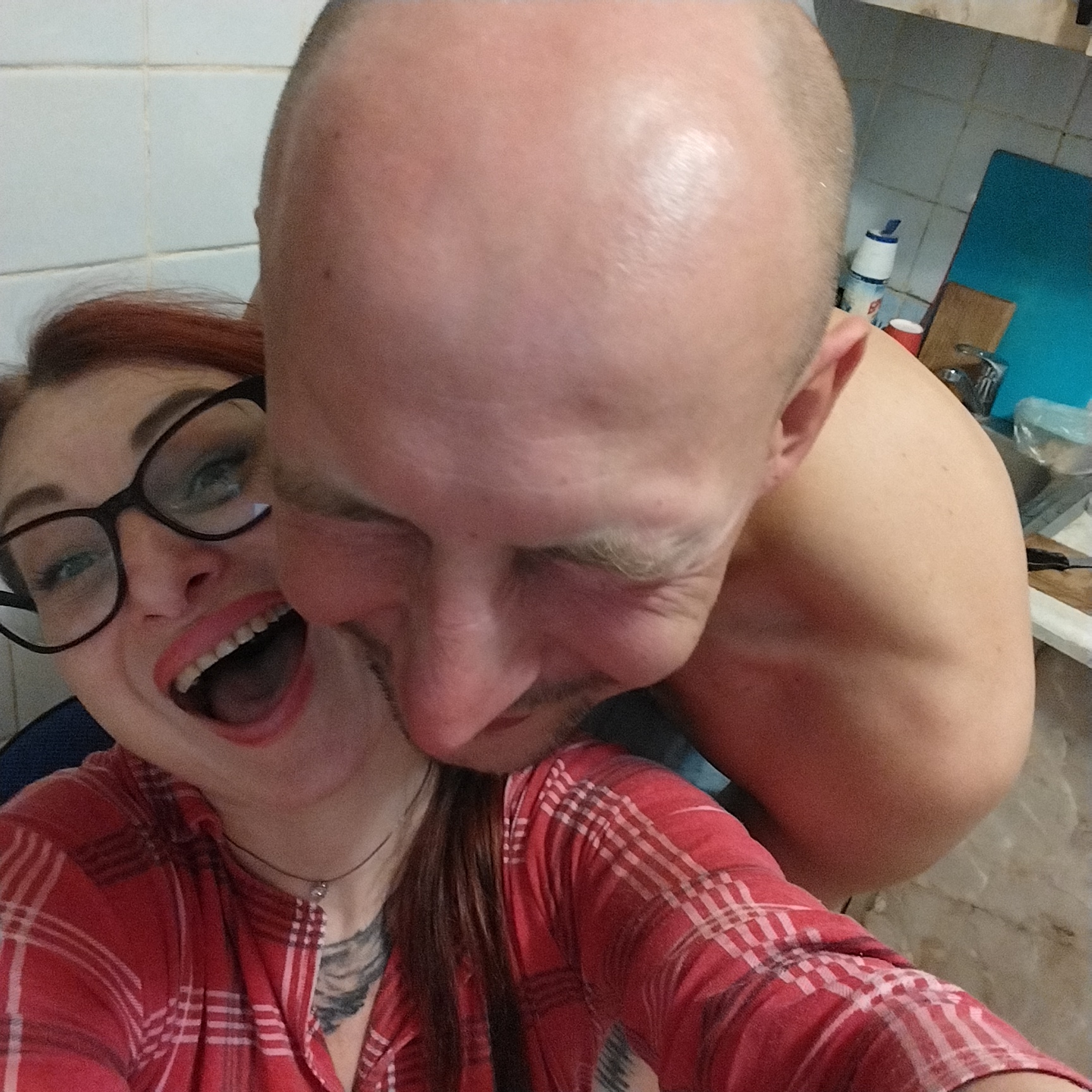 Two people are posing for a selfie. The person in the foreground on the left is a woman with red hair and glasses. She is wearing a red plaid shirt and has an open mouth and a wide smile. She is wearing a necklace with a small pendant. A tattoo is visible on her chest. The person in the background on the right is a bald man. He is leaning in towards the woman. The background appears to be a kitchen with white tiled walls. There is a blue cutting board and a faucet visible in the background. The overall lighting is bright.