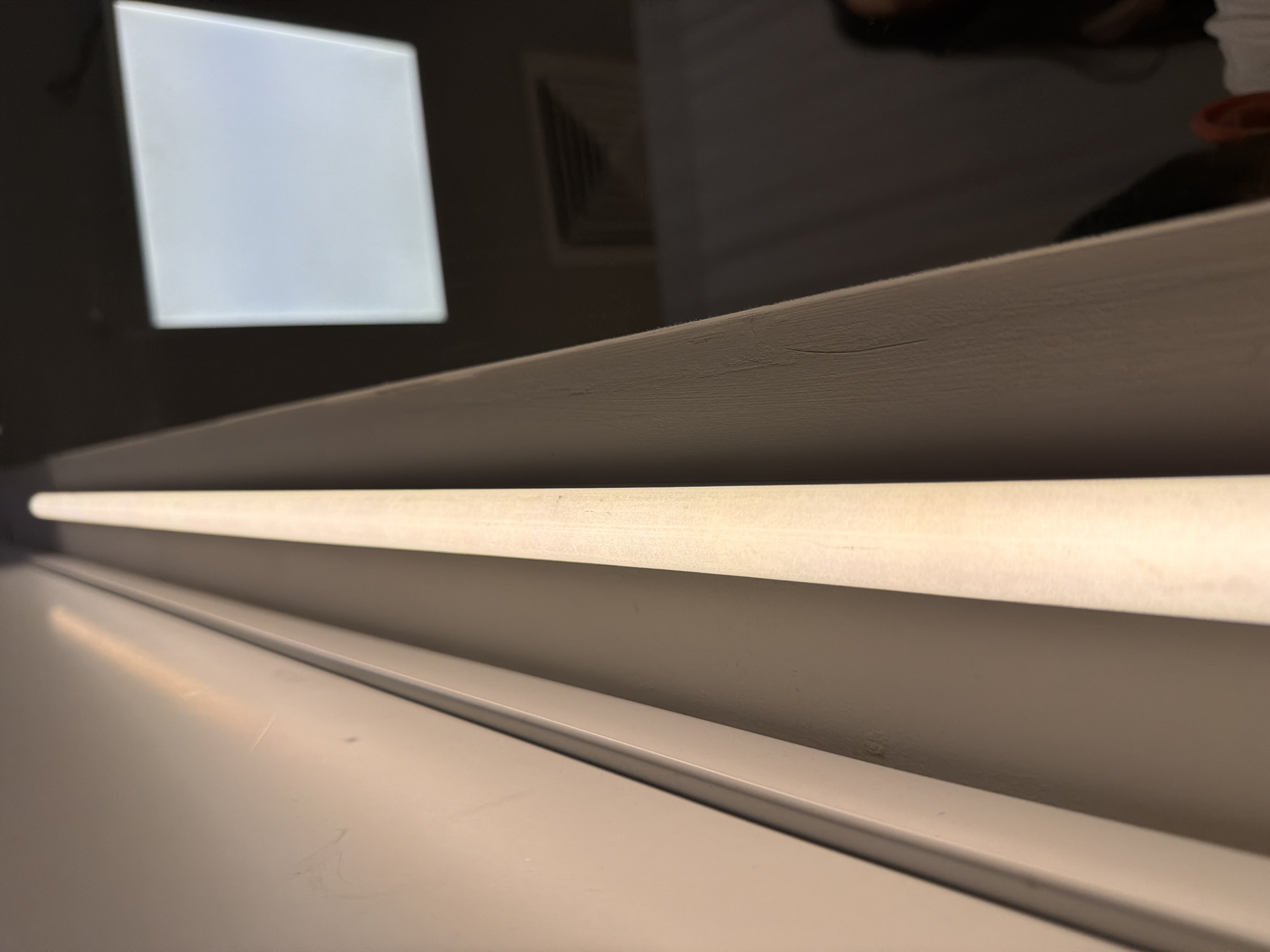 The image shows a close-up of a white shelf or ledge with a fluorescent light tube running horizontally across the upper part of the frame. The light is bright and casts a reflection on the surface below. In the background, a square white light panel is visible, likely a light fixture or a screen. The setting appears to be indoors, possibly a room or a studio, given the artificial lighting and reflective surfaces. There are hints of a dark, reflective surface above the shelf, where a blurred reflection of a person's head and ear, as well as an air vent, can be seen. The overall mood is neutral and the image is likely an abstract or detail shot focusing on light and form. There is no visible text in the image. The location context provided, Izhevsk, Russia, does not offer any specific visual cues within the image itself.