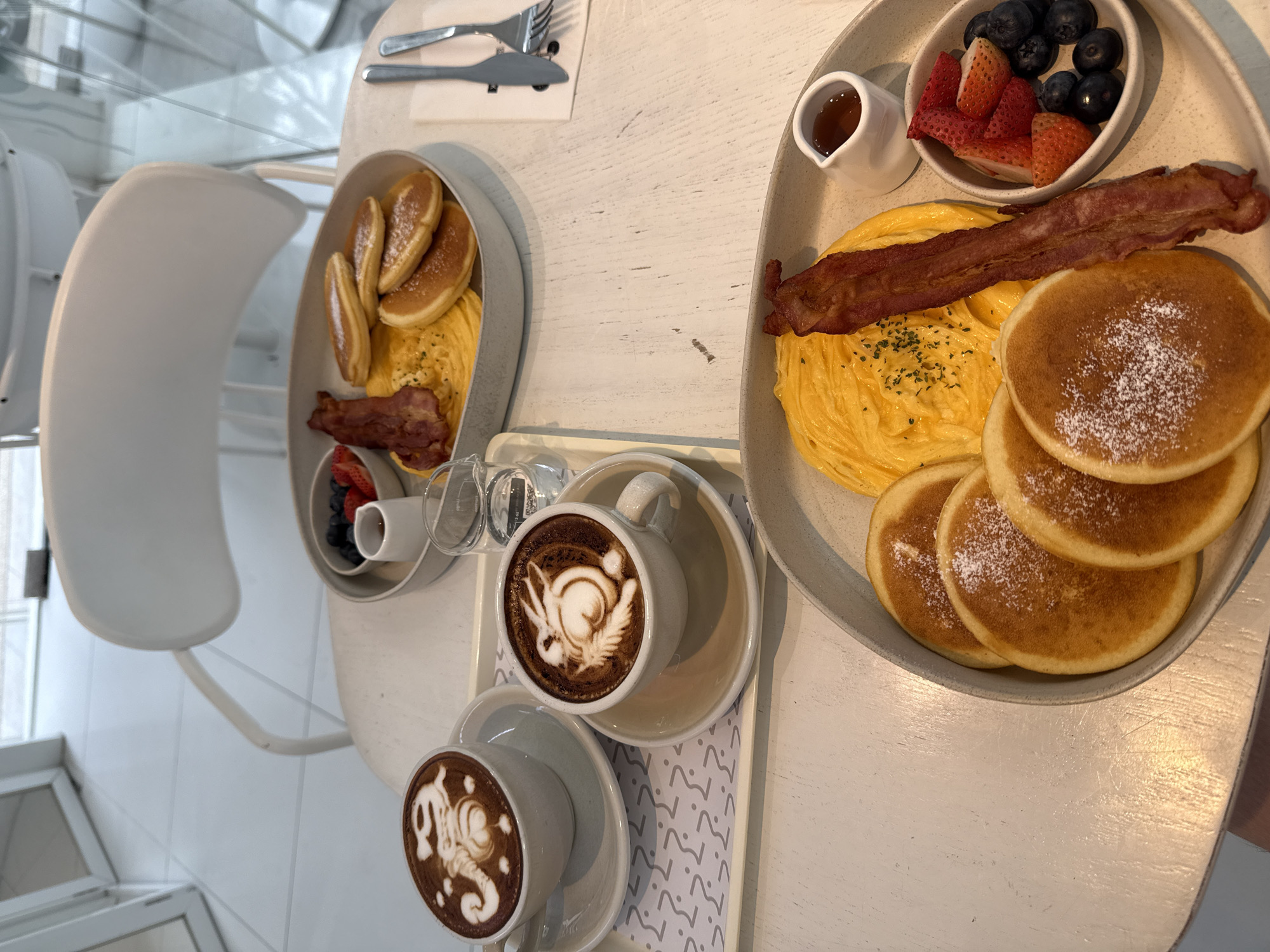 This is a close-up, high-angle shot of two breakfast plates and two cups of coffee on a white table. The setting appears to be an indoor cafe or restaurant, likely in Bangkok, Thailand, given the presence of latte art on the coffee cups and the overall aesthetic.
The plate in the foreground contains four fluffy pancakes dusted with powdered sugar, a strip of crispy bacon, a serving of fluffy scrambled eggs, a small dish of syrup, and a bowl of fresh berries including blueberries and sliced strawberries. The plate in the background has three pancakes, scrambled eggs, a strip of bacon, and a small dish of berries with a small ramekin of syrup.
Two white ceramic mugs with saucers are on a white tray to the left of the foreground plate. Each mug contains coffee with intricate latte art, one appearing to be a rabbit or animal, and the other a similar stylized design. A glass of water is also on the tray.
The table is white and appears to be made of wood with visible grain. In the background, a white chair with a modern design is visible, along with what looks like a large window and reflective surfaces, suggesting a contemporary cafe setting. There are no people clearly visible in the frame, but the setup implies two people are being served breakfast. The lighting is bright, indicating it is daytime.