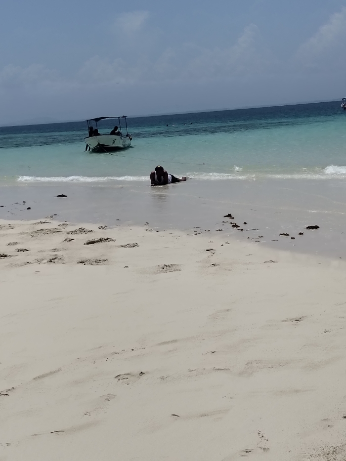 The image captures a sunny coastal scene in Wasini, Kenya. A single individual, clad in dark bottoms and a light-colored top, lies prone at the water's edge on a white sandy beach, with their lower body partially submerged in the shallow, light blue ocean water. Their head is resting on their forearms. Gentle waves break immediately in front of them. In the mid-distance, a white motorboat with a dark canopy is stationary in the clear, light blue water, with two indistinct human figures visible aboard. The water transitions to a deeper blue further from the shore, meeting a distant horizon under a light blue sky with scattered white clouds. Dark organic debris is present on the sand near the waterline, and footprints are discernible on the foreground sand.