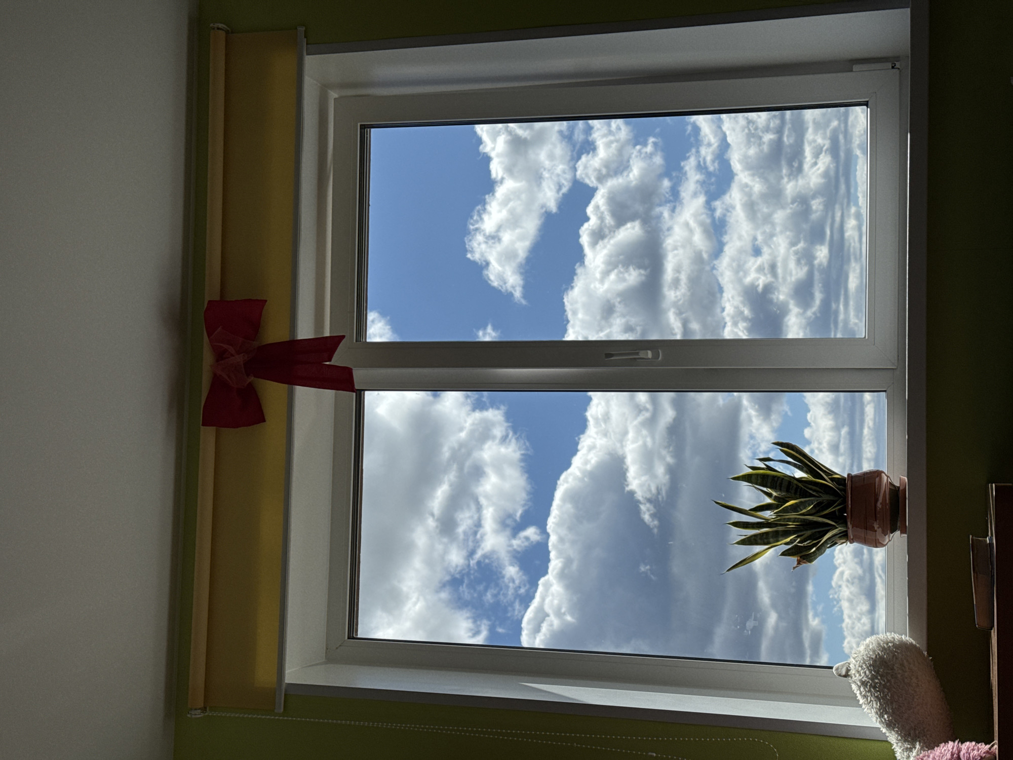 The image shows a view through a white-framed double-paned window. Outside the window, there is a clear blue sky with fluffy white clouds. A potted snake plant sits on the windowsill. A yellow window shade is partially rolled up on the left side of the window, and a red ribbon is attached to it. A stuffed animal is placed on the windowsill in the lower right corner. The walls are painted in two colors: a light green on the lower portion and white on the upper portion. The location is Прикамский, Russia.