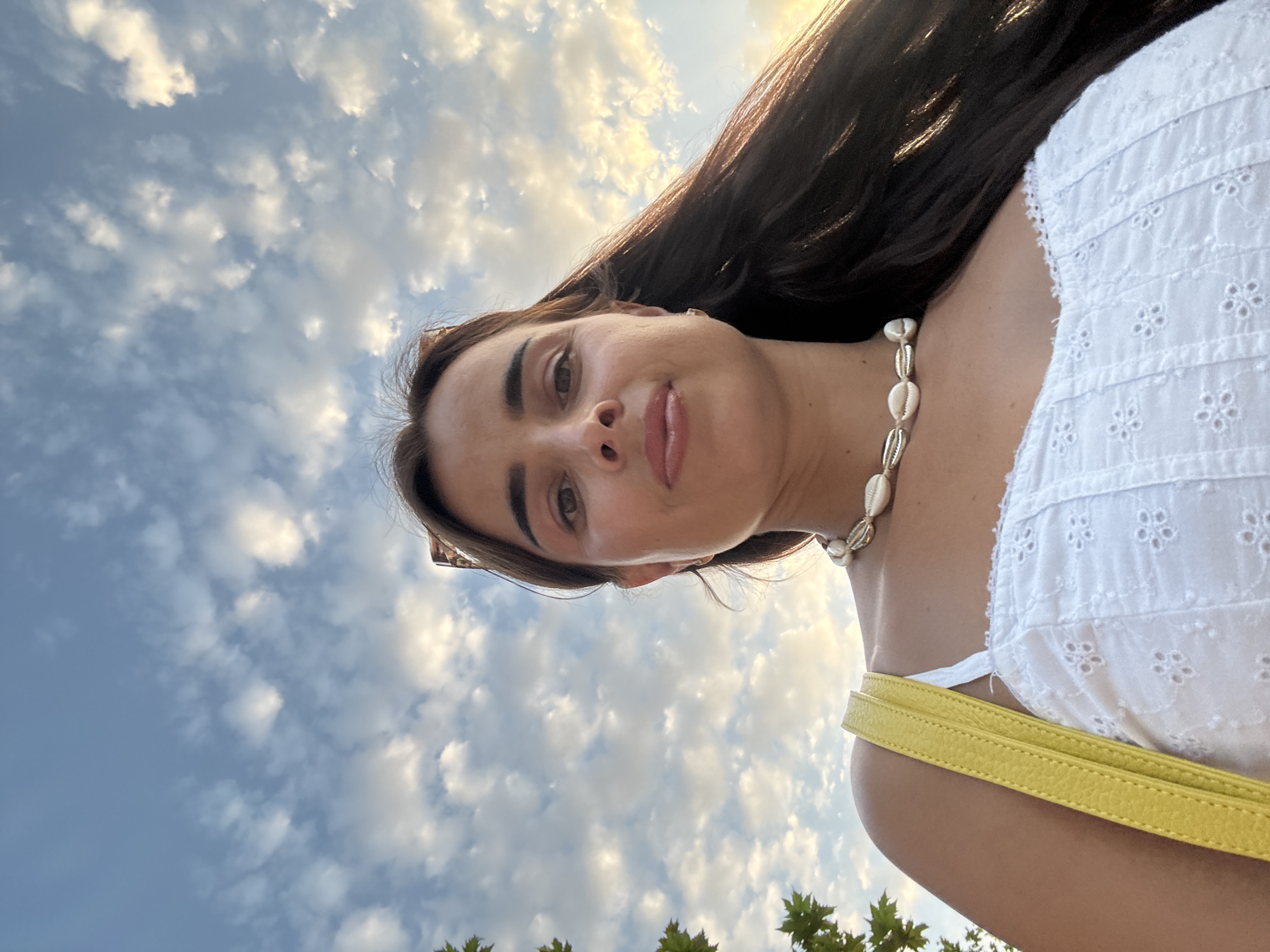 A woman is taking a selfie outdoors with a cloudy sky in the background. She has long dark hair, and is wearing a white eyelet top and a shell necklace. A yellow purse strap is visible on her left shoulder. The lighting suggests it is either sunrise or sunset.