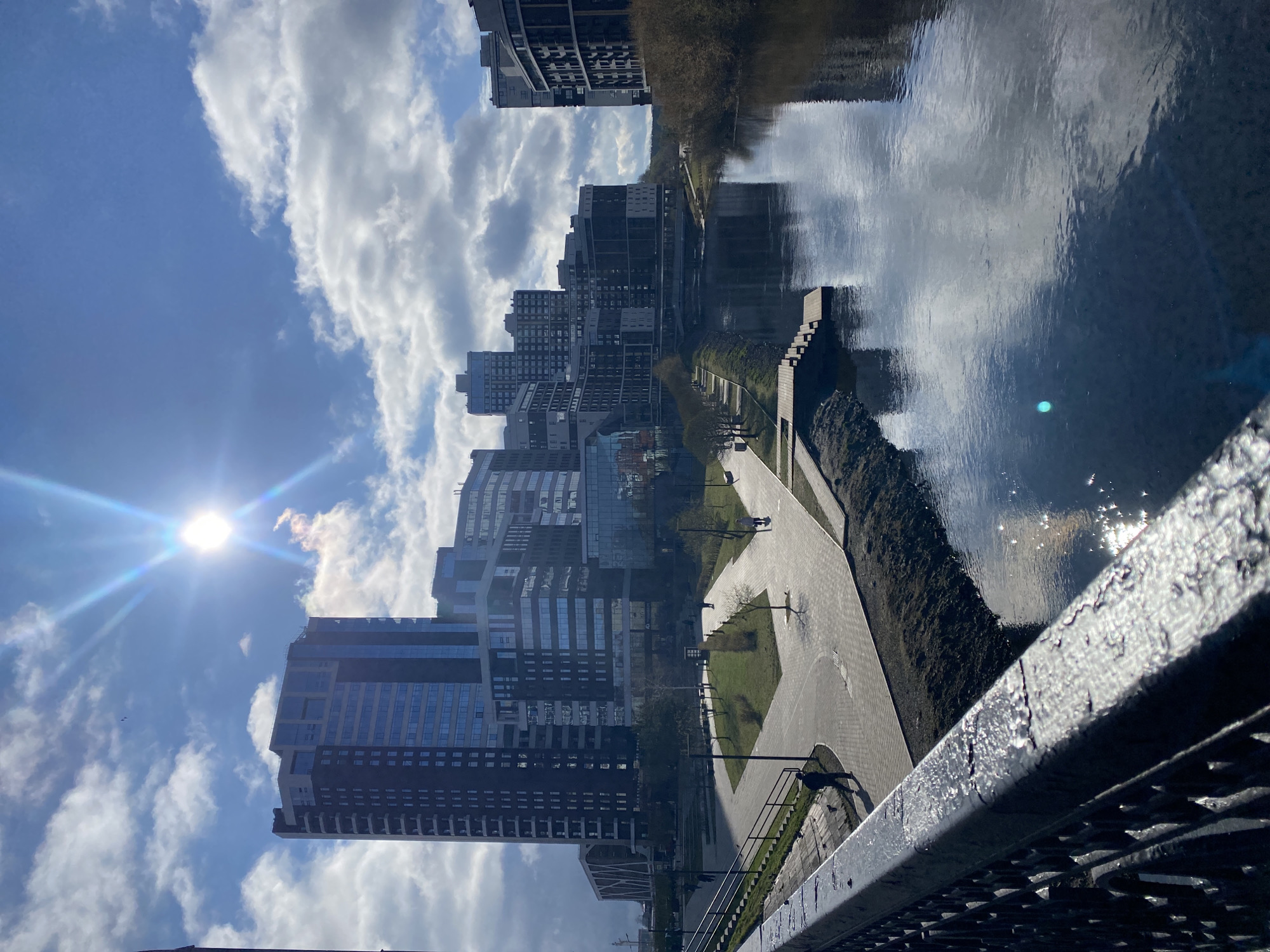 This is a bright, sunny daytime photograph depicting an urban waterfront scene, likely in Yekaterinburg, Russia, taken from an elevated vantage point, possibly a bridge. The original image is rotated 90 degrees counter-clockwise.
The sky, occupying the upper portion of the corrected frame, is a vibrant blue with scattered white cumulus clouds. The sun shines intensely from the upper left, creating a noticeable lens flare. Modern, multi-story buildings with dark facades and large reflective glass windows line the left side, mirroring the sky and clouds.
A wide, calm river or canal dominates the right side of the image, serving as a large natural mirror that perfectly reflects the sky, clouds, and the distant cityscape. The sun's glare creates sparkling highlights on the water's surface.
Along the riverbank on the left, a paved pedestrian pathway stretches into the distance, flanked by patches of green grass and sparsely leafed trees, suggesting late autumn, winter, or early spring. Several small figures, appearing as pedestrians, can be seen on this pathway. Some steps leading down to the water are also visible.
In the immediate foreground on the lower right, a dark, textured metal railing with chipped paint provides a sense of the observer's position on a structure overlooking the scene. The overall atmosphere conveys a clear and pleasant day in a modern city environment. No visible text is present in the image.