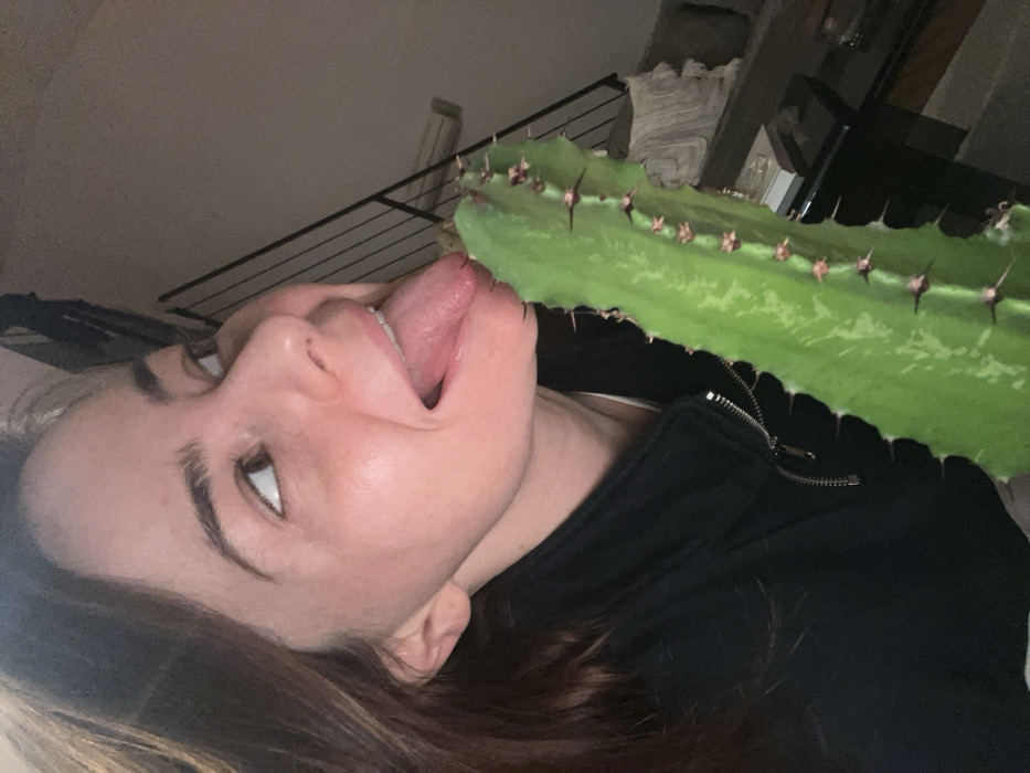 A woman with brown hair and fair skin has her tongue extended towards a tall, green cactus with prominent spines. She is looking up and to the left with her mouth open. She wears a black zipped-up jacket. The background is dimly lit, with glimpses of a staircase railing and furniture. The image is taken at an angle, making the scene tilted.