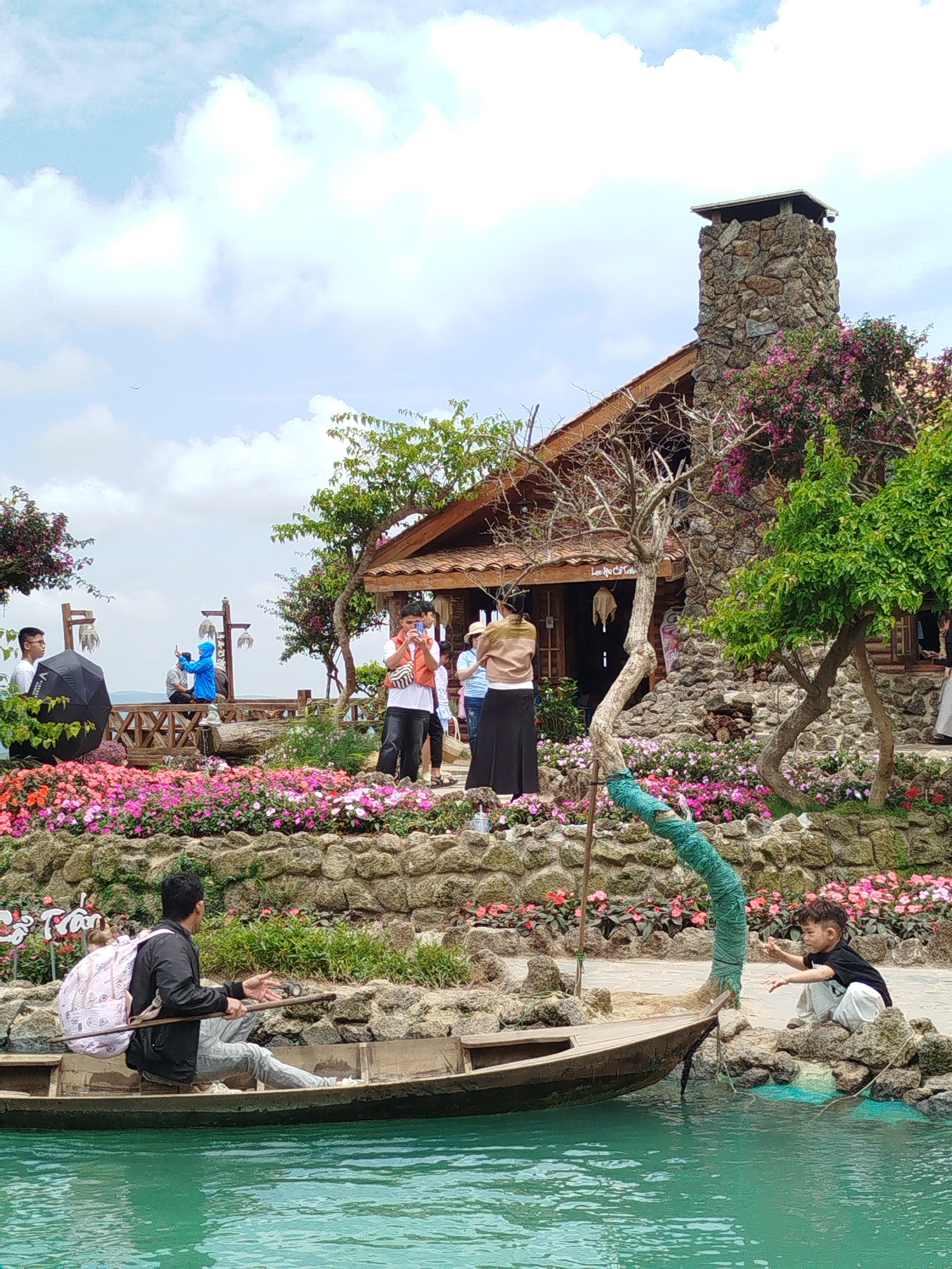 This is a full outdoor, eye-level shot of a scenic location in Xuân Thành, Vietnam, likely a tourist attraction, featuring a traditional-style wooden house with a prominent stone chimney. In the foreground, a man with a pink backpack sits in a small wooden boat on turquoise water, holding a paddle. To the right, a young child in white pants and a black t-shirt sits on rocks near the water's edge. Behind the boat, a lush garden bursts with colorful pink and red flowers cascading down stone walls. A stone path winds past the flowers, leading towards the house. Several people are visible on a wooden deck in the midground, some taking photos. The house itself is adorned with flowering vines, and trees with green foliage surround it. The sky is partly cloudy with a light blue hue.