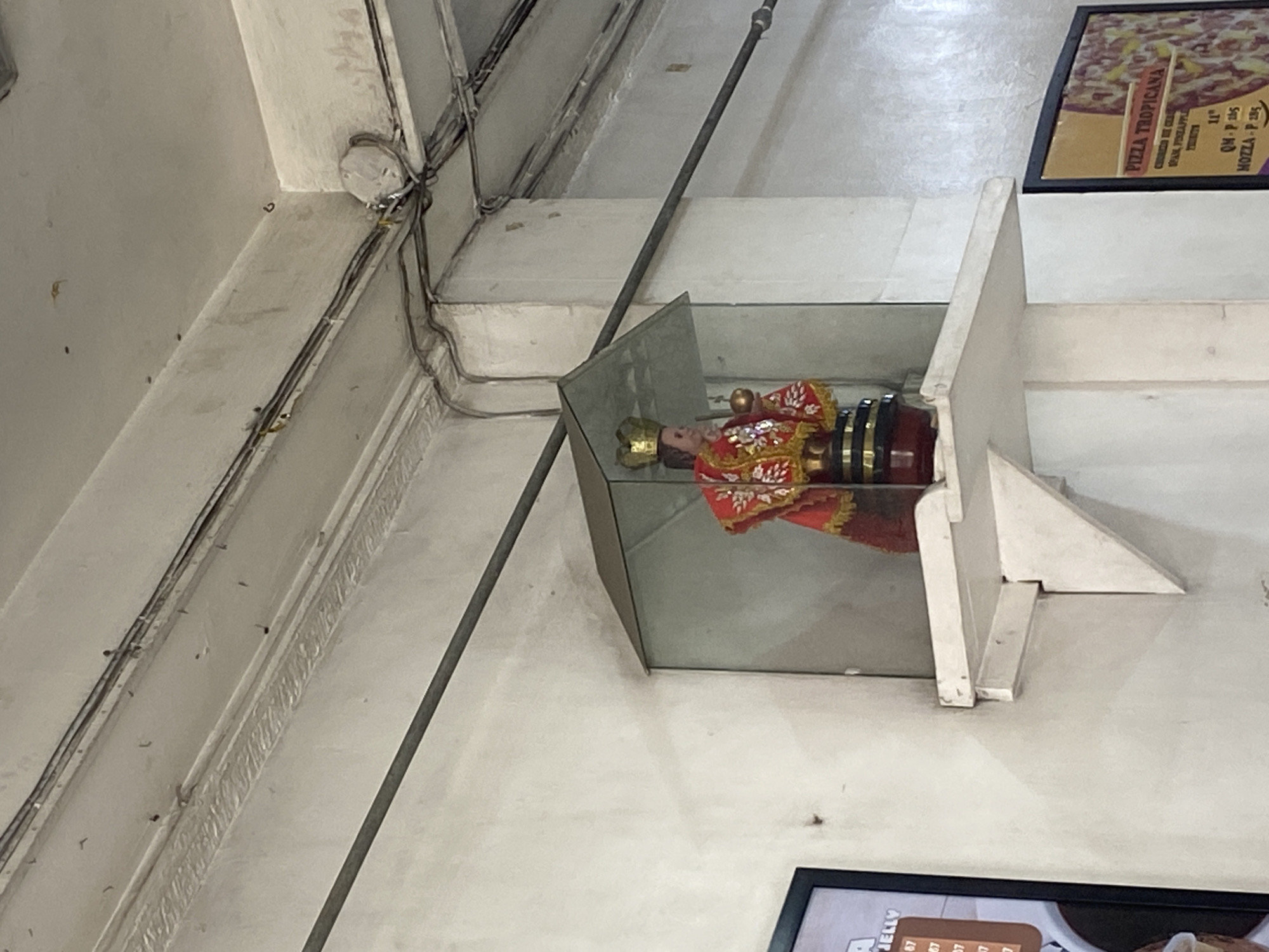 This interior image, captured in Davao City, Philippines, depicts a section of a commercial establishment, likely a restaurant or canteen, with a distinctly Filipino aesthetic. The central focus is a small, ornate religious statue, resembling the Santo Niño (Infant Jesus), encased in a glass display box. The figure wears a gold crown and elaborate red and gold robes, resting on a black and gold base, positioned on a white, angular shelf.
Above and to the left of the statue, exposed electrical conduits and wiring run along the concrete ceiling and beams, showcasing a utilitarian installation. A long, thin metal pole extends diagonally across the frame, partially obscuring the background.
Two framed signs, presumably menu boards, are visible on the white walls. The upper right sign, tilted with the image, advertises "PIZZA TROPICANA" with details such as "SPAM, PINEAPPLE" as ingredients and "THICK" crust. Prices are listed in Philippine Pesos: "QM - P 265" and "MOZZA - P 285." A partially visible sign in the lower right corner shows the word "JELLY" followed by the number "67," likely indicating a dessert or beverage option and its price. The scene is brightly lit, suggesting a well-illuminated indoor space. No explicit human activity is visible.