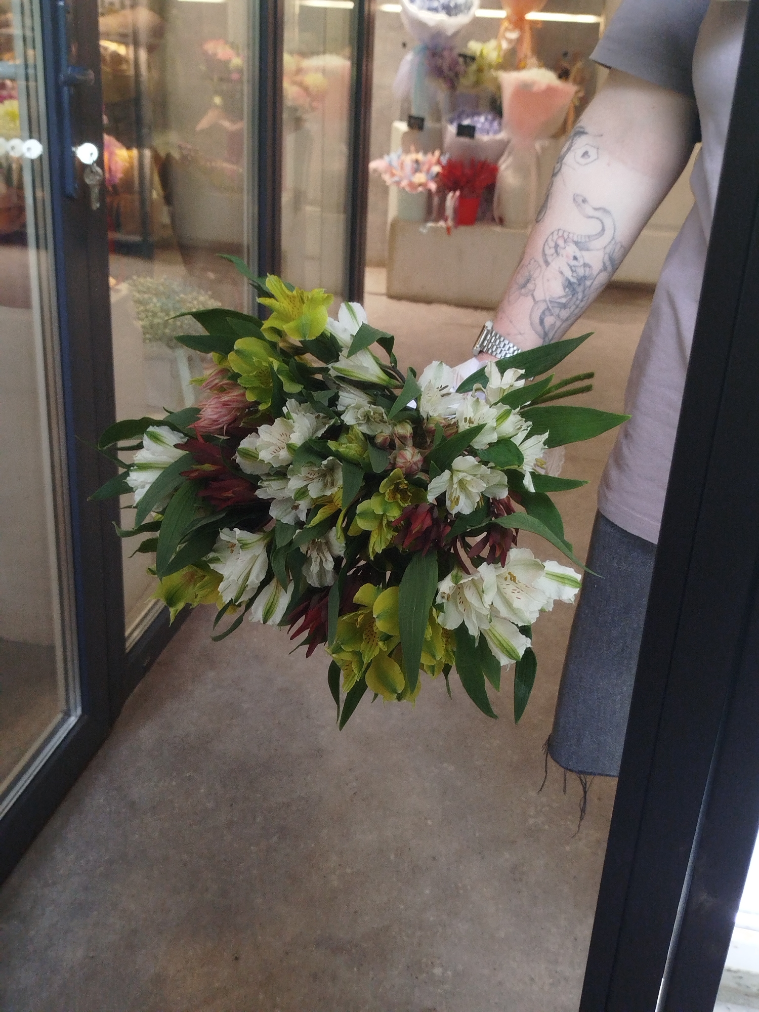 A person, partially visible, holds a bouquet of white and green alstroemeria flowers in front of a glass-paned doorway. The person is wearing a gray t-shirt and a dark gray apron. Their left arm, which is tattooed with a snake and other designs, is visible holding the bouquet. The background shows a glimpse of a flower shop with various floral arrangements and a key in the lock of the door. The floor is a concrete surface. The overall setting appears to be an indoor space, possibly a retail store.