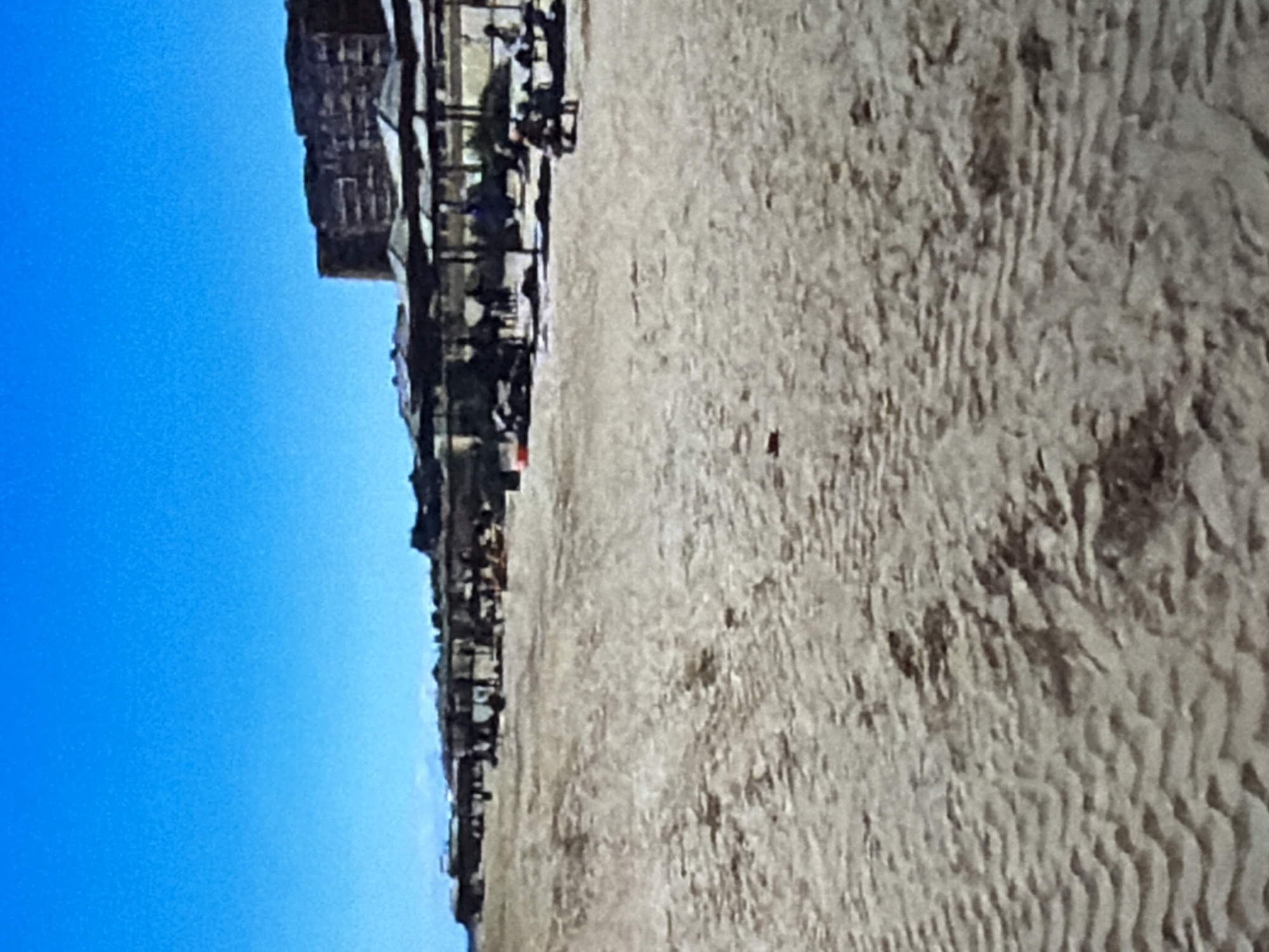 The image depicts a sandy beach with a clear blue sky. In the background, a multi-story building complex with dark facades stands against the sky. Along the edge of the beach, there is a row of beach umbrellas and chairs, suggesting a recreational area. Some people are visible in the distance, appearing as small figures enjoying the beach. The sand shows patterns from ripples, possibly from wind or waves. The overall scene suggests a sunny day at a beach resort or a similar coastal location. The image is oriented vertically, with the sky on the left and the beach on the right.