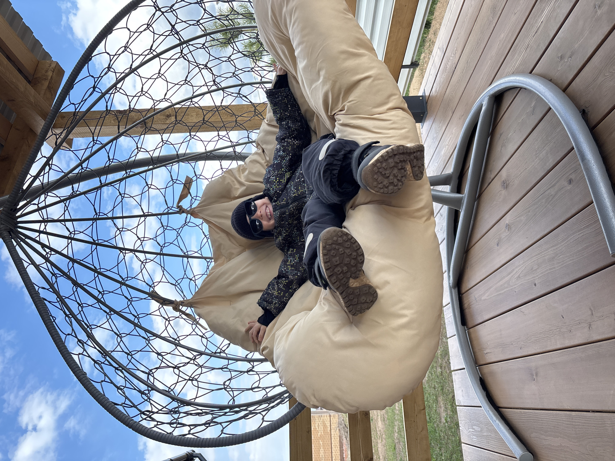 An outdoor, rotated full shot captures a young child lying comfortably in a large, round hanging chair in Chaikovsky, Russia, on a bright day. The child, appearing to be a boy, is dressed in winter attire including a dark patterned jacket, dark pants, a black beanie, and sunglasses, and wears sturdy boots. He is nestled within thick, beige cushions that line the inside of the chair, which features a prominent metal cage-like frame. The child smiles broadly towards the camera, suggesting a happy and relaxed mood.
The background reveals a clear blue sky dotted with fluffy white clouds through the intricate metalwork of the chair. To the right of the frame, a wooden deck with light brown horizontal planks is visible, along with a curved, grey metal base or support structure for the swing. Hints of green grass and distant light-colored brick buildings can be seen beyond the deck. The setting appears to be an outdoor recreational area, likely a patio or balcony. No visible text is present.