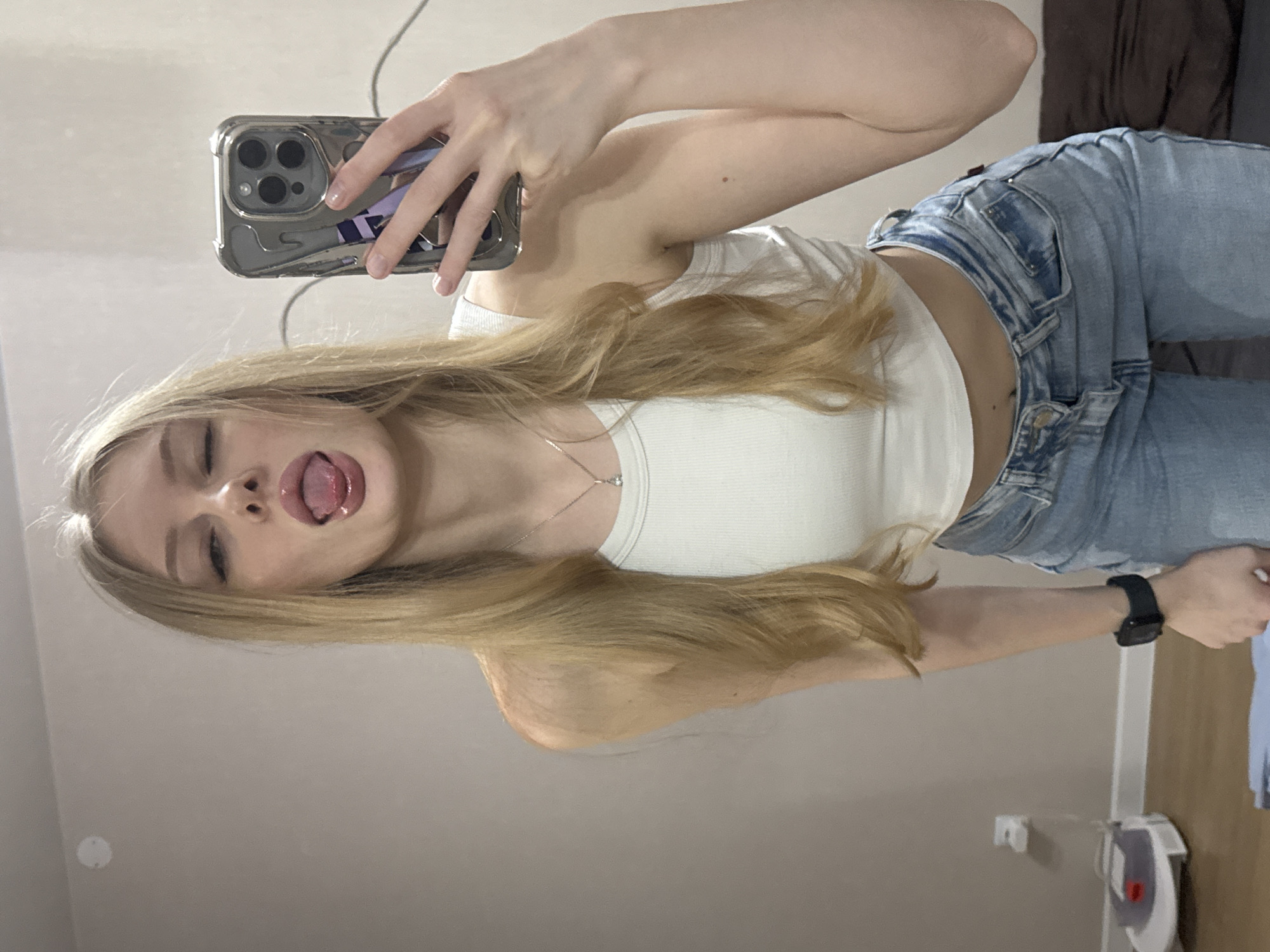 The image, taken in Perm, Russia, features a young woman capturing a selfie indoors. She has long, straight blonde hair and is depicted wearing a white ribbed tank top and light blue denim jeans. Her right arm is raised, holding a smartphone with a triple-lens camera array and a metallic-looking protective case, which shows a hint of purple design. A dark smartwatch is visible on her left wrist, and she wears a silver pendant necklace.
The woman's face is central, displaying a playful expression with her eyes partially closed and her tongue playfully sticking out. The background is simple, consisting primarily of a light-colored wall. In the upper right corner, a dark brown object, possibly furniture like a headboard or couch, is partially visible. The lower right reveals a light-colored vertical structure and a small white appliance with a red dial. A cable or wire can be seen hanging in the upper left quadrant. The scene conveys a casual and lighthearted moment.