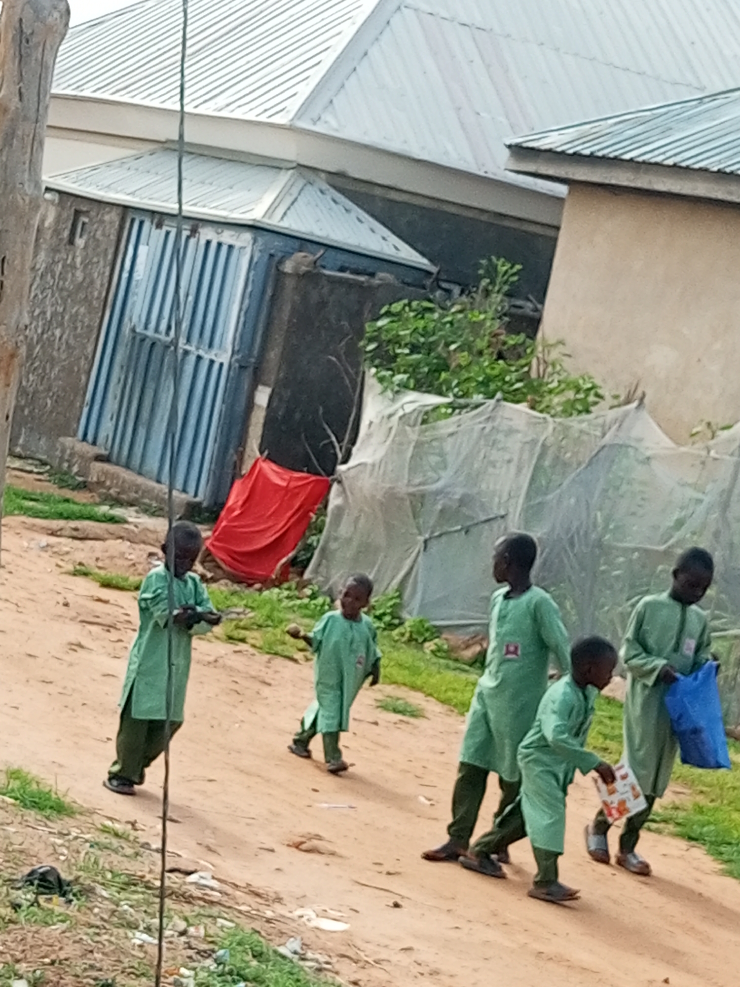 In Tempure, Nigeria, a group of five young boys, all wearing matching green uniforms, walk along a dirt path. The boy in the front on the left is holding a small book. The boy in the center is looking down as he walks. The boy in the middle on the right is holding a blue plastic bag. The boy in the back on the right is also carrying a blue plastic bag. In the background, there are several buildings with corrugated metal roofs, one of which is painted a tan color. There are also some leafy green plants visible. The ground is covered in loose dirt and small rocks.