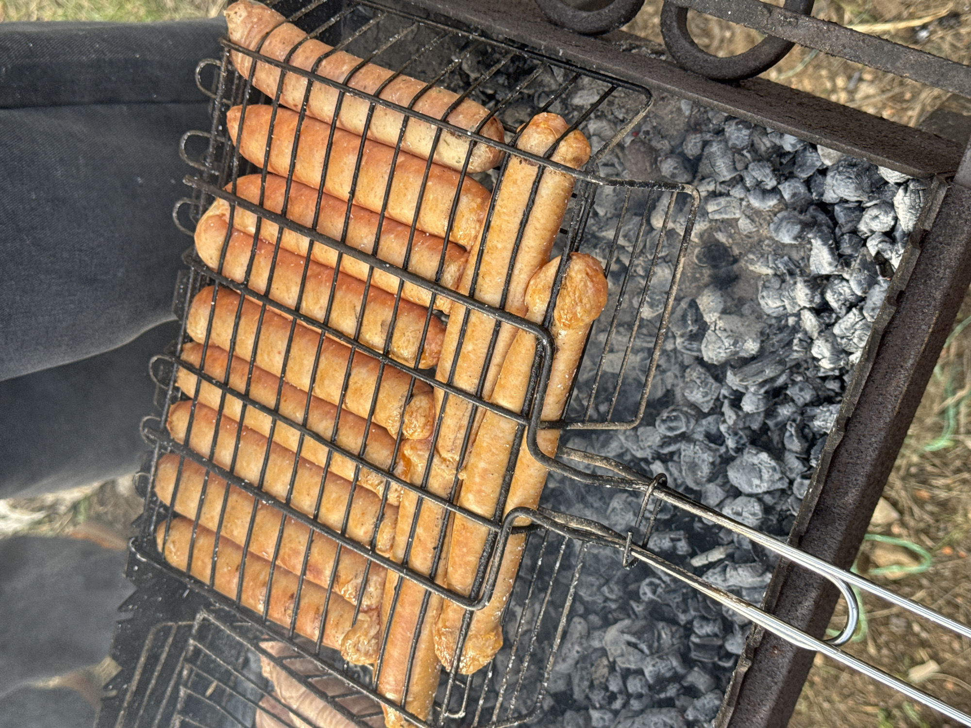 A close-up, high-angle shot depicts sausages grilling in a wire basket over a charcoal grill. The sausages appear lightly browned and are arranged neatly within the confines of the basket. The charcoal underneath is mostly gray ash, indicating that the fire is well established. The grill itself appears to be made of dark metal, and the handles of the basket extend toward the right side of the frame. A person wearing dark clothing is partially visible in the upper-left corner of the frame. The presence of what appears to be grass and soil in the background suggests that the grilling is occurring outdoors. There is no indication of the location being Abrosy, Russia, or any urban setting.