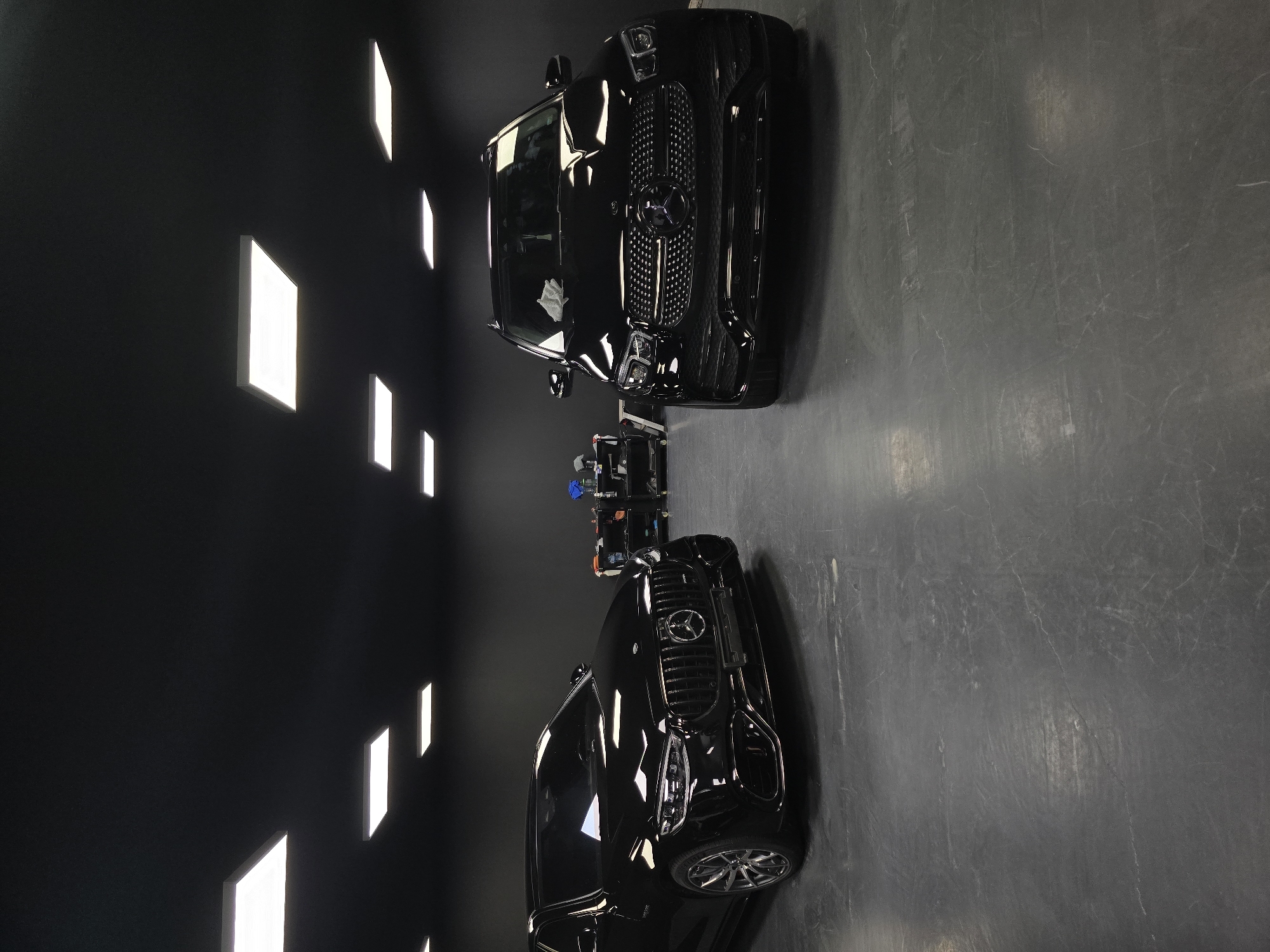 In a dimly lit, black-walled garage or detailing studio, two high-end black Mercedes-Benz vehicles are parked side-by-side. The car on the left appears to be a sedan, possibly an S-Class, with its front grille prominently displayed. The car on the right is an SUV, likely an AMG GLE. Both vehicles are clean and shiny, reflecting the overhead square LED panel lights that are recessed into the black ceiling. In the background, a multi-tiered rolling cart laden with car detailing supplies and equipment is visible, suggesting a professional automotive service environment. The floor is a dark, concrete-like surface with visible scuff marks and wear. There are no people visible in the image. The scene does not contain any specific elements that would indicate a location in Ruse, Bulgaria; it depicts a generic, high-quality car detailing or storage space.