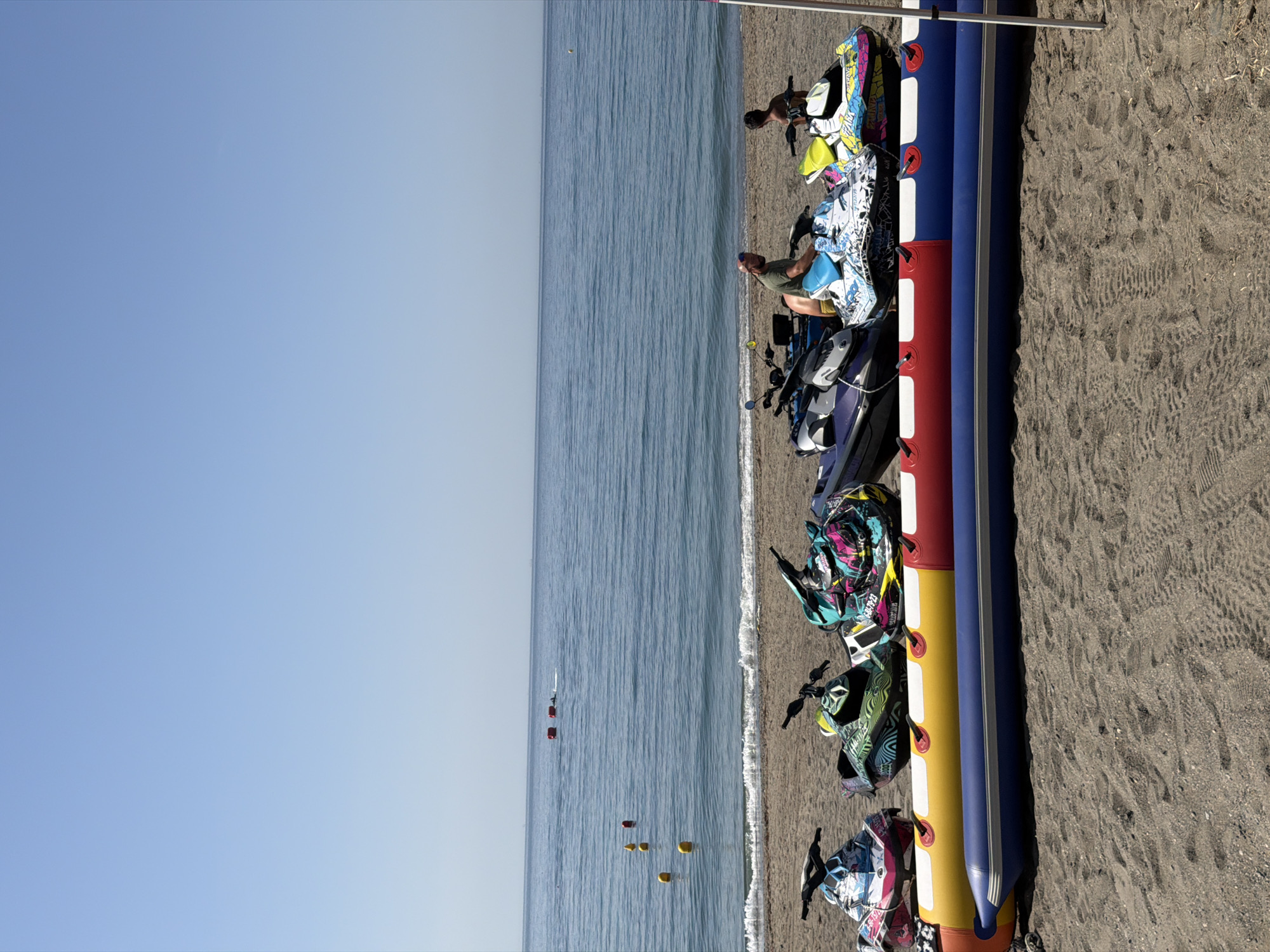 The image depicts a sunny day at a sandy beach in Estepona, Spain, with the Mediterranean Sea in the background. Lined up on the shore are several colorful jet skis, some with people on them. In front of the jet skis is a large inflatable banana boat, painted red, yellow, and blue. Two men are visible on or near the jet skis. One man, wearing a green t-shirt, is seated on a jet ski, appearing to interact with it. Another younger man, shirtless, is standing behind another jet ski. The sea is calm with gentle waves lapping the shore, and buoys are visible in the water further out. The clear blue sky suggests pleasant weather conditions.