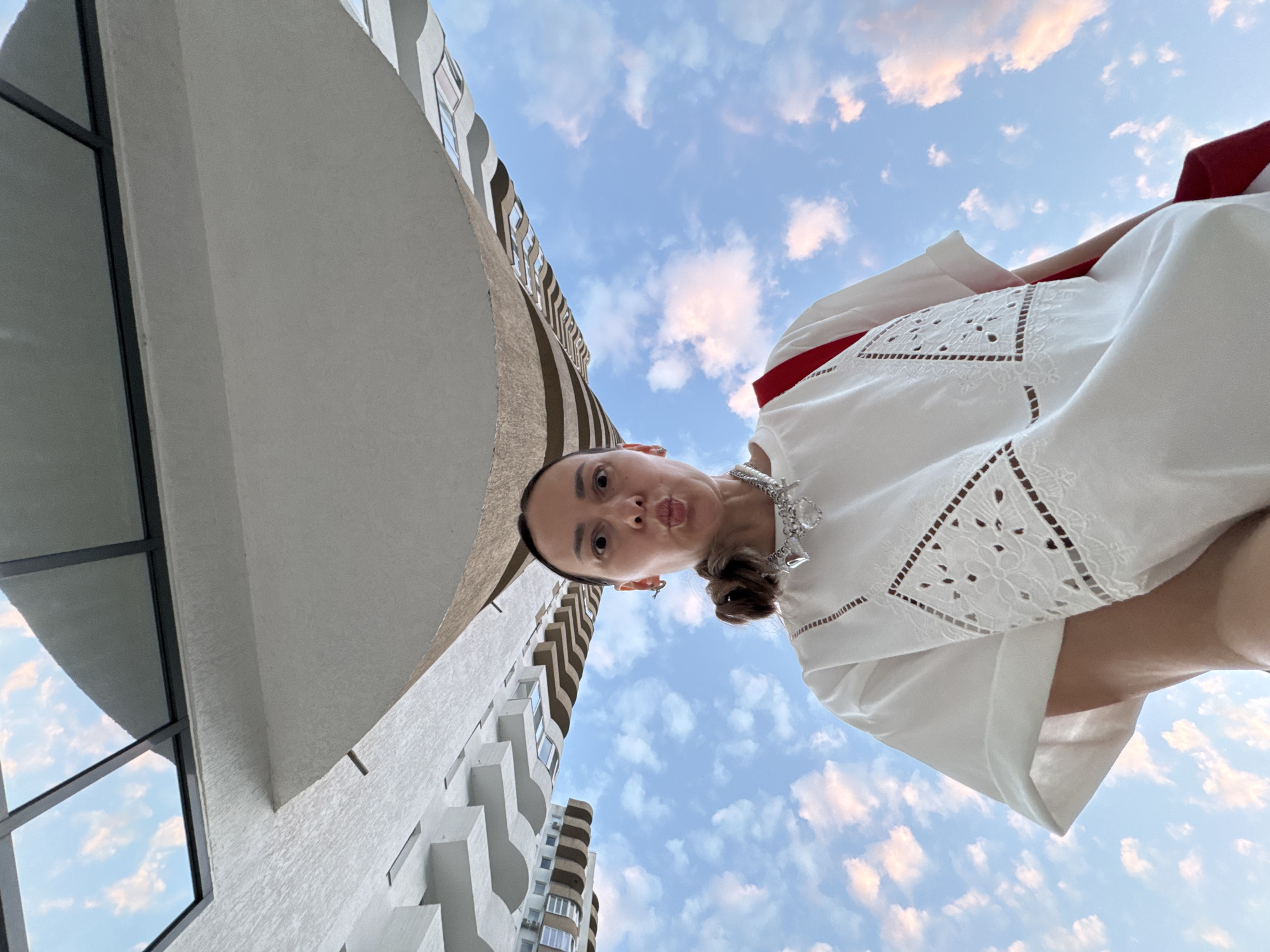 A woman is posing for a selfie with a smartphone, looking up at the camera with her lips pursed. She is wearing a white dress with intricate lace detailing on the front and a red strap across her shoulder. A silver necklace with charms, including hearts and a gemstone, adorns her neck. Her hair is pulled back into a ponytail. The background shows a wide-angle view of a modern building with balconies and a clear blue sky with scattered clouds, suggesting an outdoor urban setting. The camera angle is from below, looking upwards at the woman and the architecture. The scene is outdoors during the day, likely in a city. It is not possible to identify the location as Sochi, Russia, from the image alone.