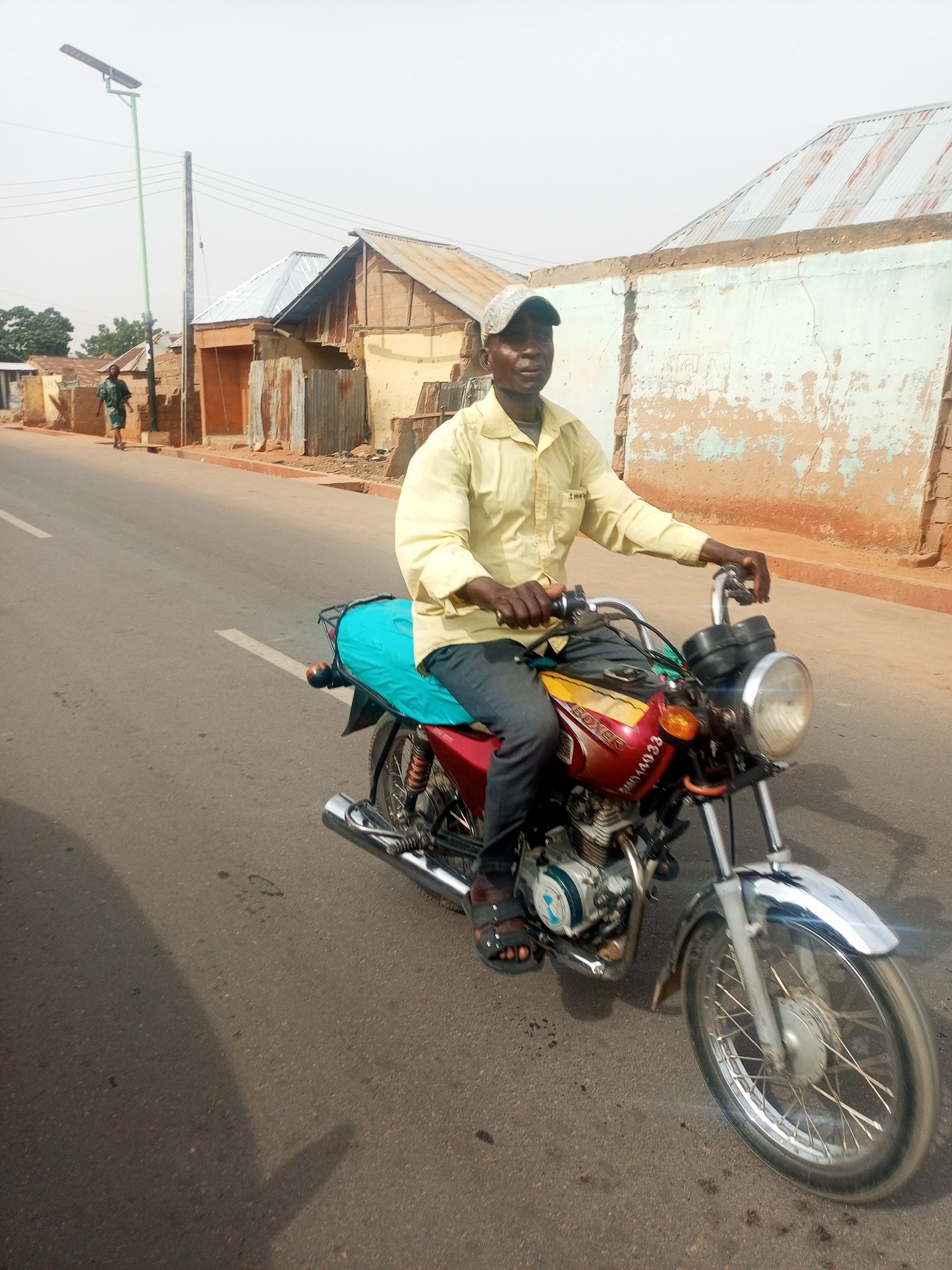 This image captures a daytime scene in Tempure, Nigeria, focusing on a man riding a red and yellow Bajaj Boxer motorcycle on an asphalt road. The man, wearing a light yellow long-sleeved shirt, dark trousers, sandals, and a light-colored baseball cap, is positioned slightly right of the frame's center, looking towards the camera with a neutral expression. His hands are on the handlebars of the motorcycle, which features a blue cover on its seat and chrome accents on the exhaust, front fender, and headlight bezel. Text "BOXER" is clearly visible on the motorcycle's fuel tank, along with a partially obscured number plate "KMAA033" (or similar).
The setting appears to be a semi-urban or rural street lined with single-story buildings made of mud bricks and corrugated metal roofs, some showing signs of wear with peeling paint in hues of light blue, brown, and yellow. On the left side of the road, a modern solar-powered streetlight stands tall on a green pole, alongside a traditional wooden utility pole with power lines. Further down the road on the left, another person in a green garment is walking. The sky is bright and hazy or slightly overcast, suggesting a dry climate. Shadows are visible on the road, indicating daylight, likely late morning or early afternoon.