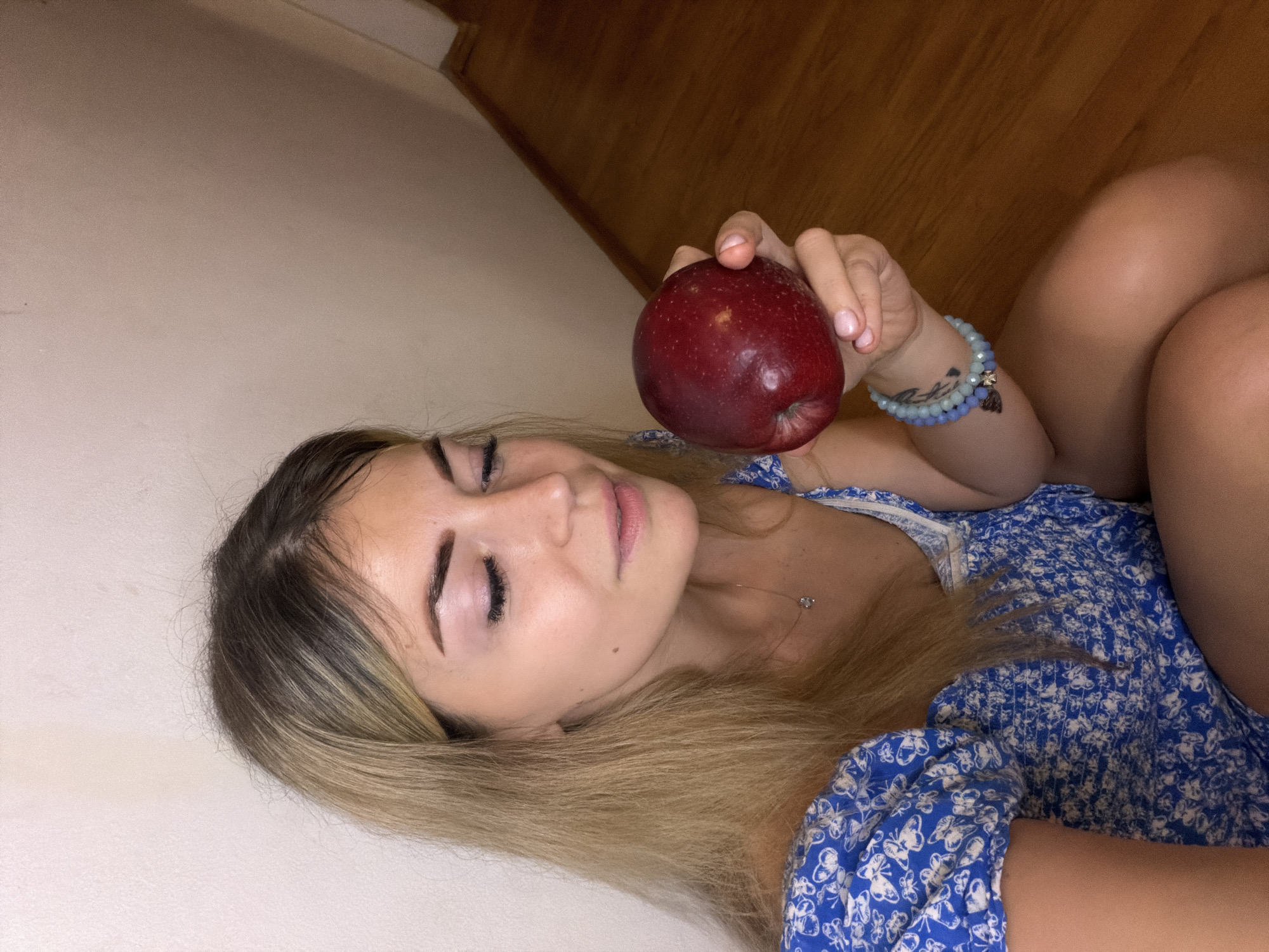This is an indoor, overhead shot of a young woman lying down, holding a dark red apple. The woman, who appears to be in her 20s or early 30s, has long, blonde hair with darker roots, fanned out around her head. Her eyes are gently closed or looking downwards, showcasing long eyelashes and neatly sculpted eyebrows. Her lips are slightly parted in a relaxed expression.
She is dressed in a blue sundress featuring a white butterfly or floral pattern. Her right arm is bent, and she is holding a large, glossy red apple prominently in the center of the frame. On her right wrist, she wears several beaded bracelets in shades of light blue and white, along with a small, dark charm and a subtle tattoo on her forearm. Her fingernails are painted with a light pink or nude polish. Parts of her bare legs or thighs are visible in the lower right corner, indicating a relaxed, reclining posture.
The setting appears to be an indoor space. The background on the left side of the image is a plain, light-colored wall, while the right side features a warm-toned wooden surface, likely a floor or wall paneling. The lighting is soft and even, suggesting an artificial or ambient indoor light. The woman's expression conveys a sense of serenity or contemplation. No specific time of day or weather can be discerned from the image, and there is no visible text. The provided location context is Budva, Montenegro, though no visual cues within the image confirm this specific location.