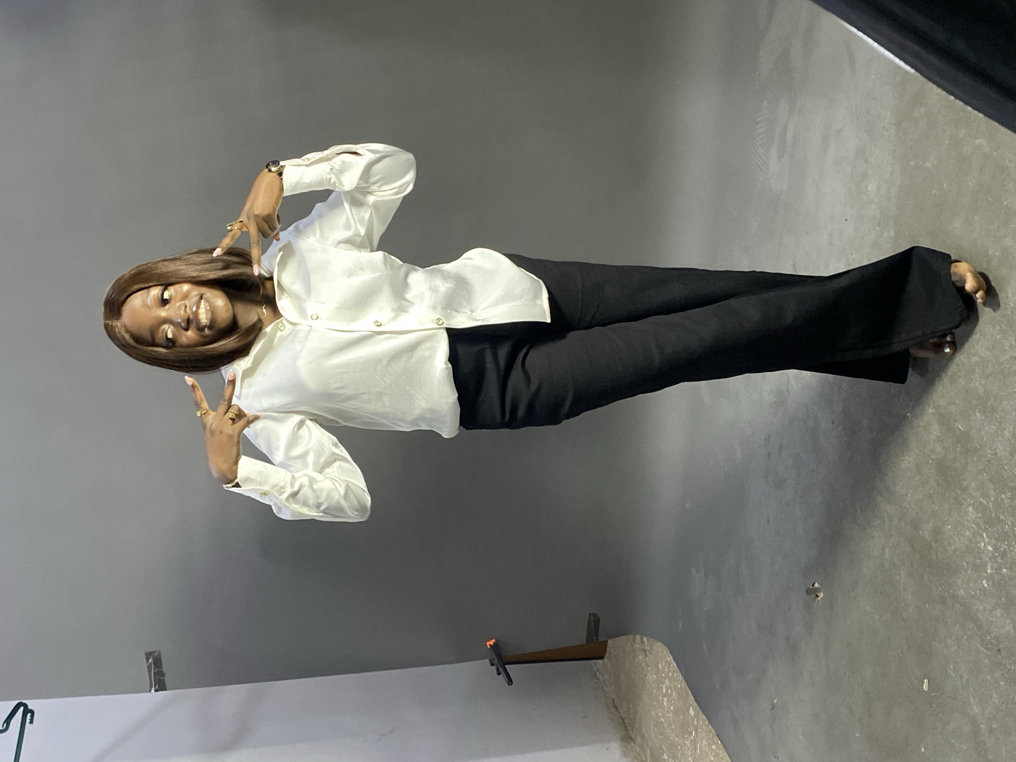A woman stands in front of a gray background. She wears a white button-down shirt and black pants, and she holds up two fingers on each hand. The background is a gray wall, and the floor is a gray concrete surface. A partial outline of a light box can be seen in the top right corner. A wall and baseboard are visible in the lower left corner. There is a brown object sticking out from the wall. The location is likely a photography studio, but the city of Jos, Nigeria, cannot be determined from the image.