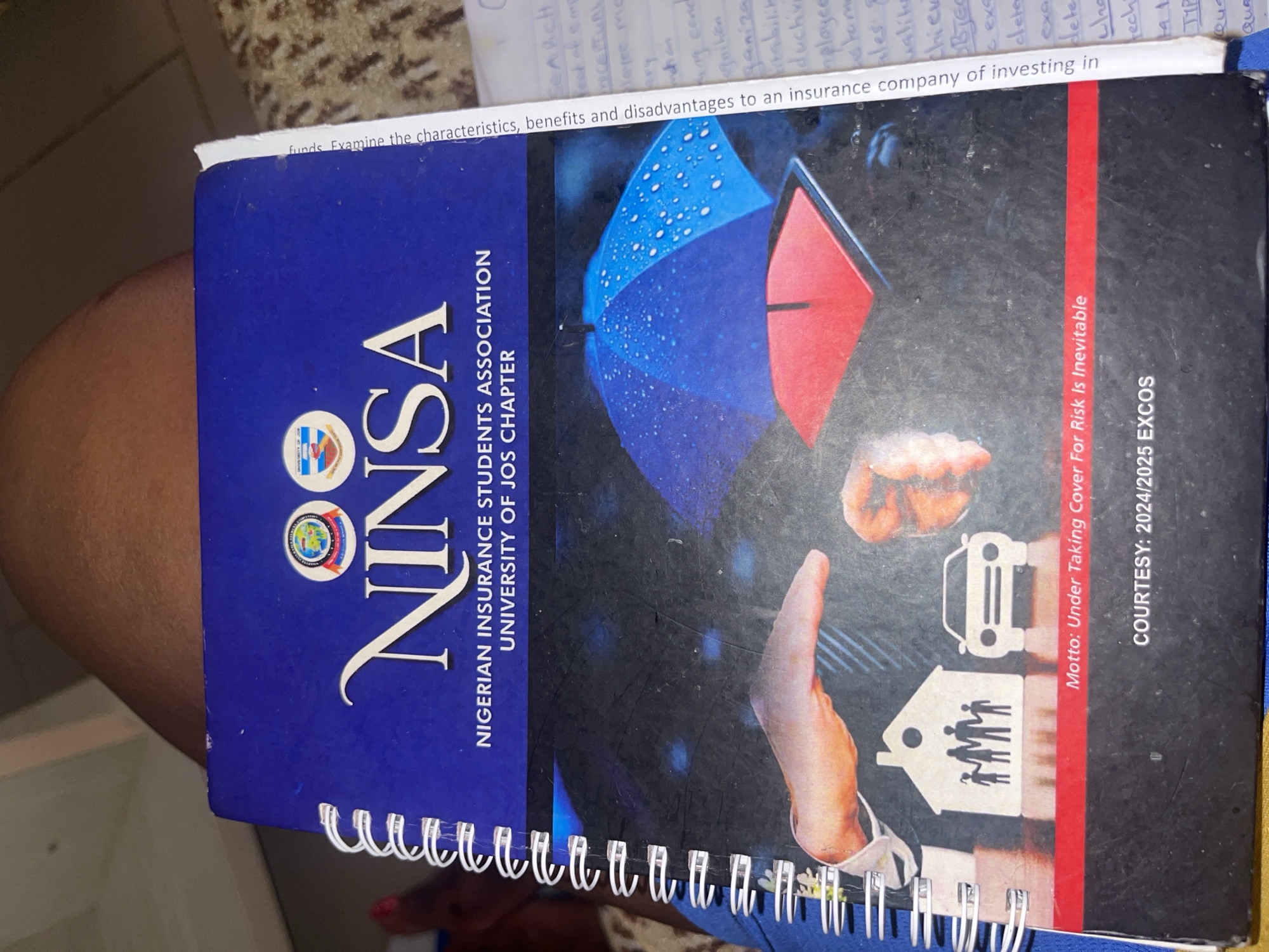 The image shows the cover of a notebook belonging to the Nigerian Insurance Students Association, University of Jos Chapter. The cover features the association's logo, the acronym "NINSA" in large white font, and the text "NIGERIAN INSURANCE STUDENTS ASSOCIATION UNIVERSITY OF JOS CHAPTER". The lower half of the cover depicts a graphic of hands holding umbrellas over silhouettes of a house with a family and a car, symbolizing protection and insurance. The motto "Under Taking Cover For Risk Is Inevitable" and "COURTESY: 2024/2025 EXCOS" are also visible. A handwritten note is partially visible on the right edge of the image, with text that appears to be related to insurance. The notebook is open and resting on a person's lap, suggesting it is in use. The background is blurred and shows a patterned surface. There is no indication of Zawan, Nigeria in the image.