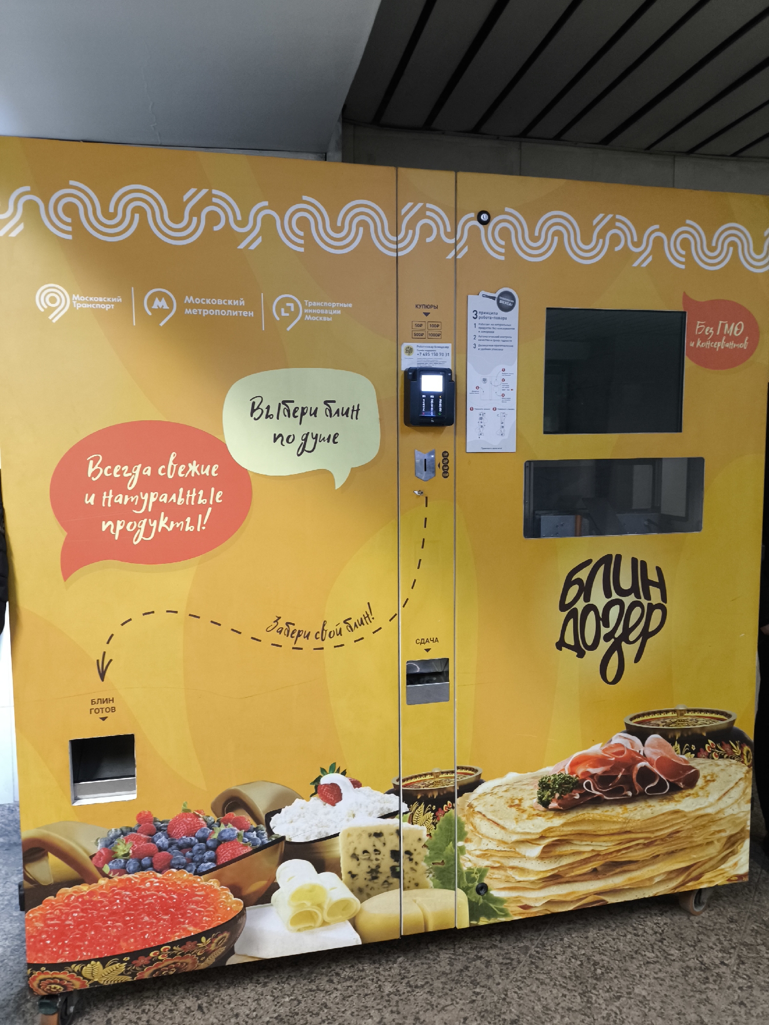 A large, bright yellow automated pancake (blini) vending machine, branded "БЛИН ДОЗЕР" (Blin Dozer), stands prominently indoors, likely within a Moscow public transport environment, as indicated by the "Московский Транспорт," "Московский метрополитен," and "Транспортные инновации Москвы" logos. The machine's surface is decorated with a warm, inviting color palette, featuring white wavy lines at the top and vibrant illustrations of blini with ham, red caviar, fresh berries, various cheeses, and butter at the bottom.
The machine features an interactive payment area with a card reader/terminal, a slot for banknotes (accepting 50P, 100P, 500P, 1000P), and a coin return labeled "СДАЧА" (Change). Two display screens are visible, along with a product dispensing slot marked "БЛИН готов" (Pancake ready). Speech bubbles convey messages in Russian: "Всегда свежие и натуральные продукты!" (Always fresh and natural products!), "Выбери блин по душе" (Choose a pancake to your liking), and "Забери свой блин!" (Take your pancake!). Another bubble declares "Без ГМО и консервантов" (Without GMO and preservatives). A small panel describes "3 принципа робота-повара" (3 principles of the robot-chef), implying an automated cooking process. A contact number, +7 495 150 70 31, is also present. The scene is well-lit, and the machine appears clean and functional, awaiting a customer. No people are visible in the image.