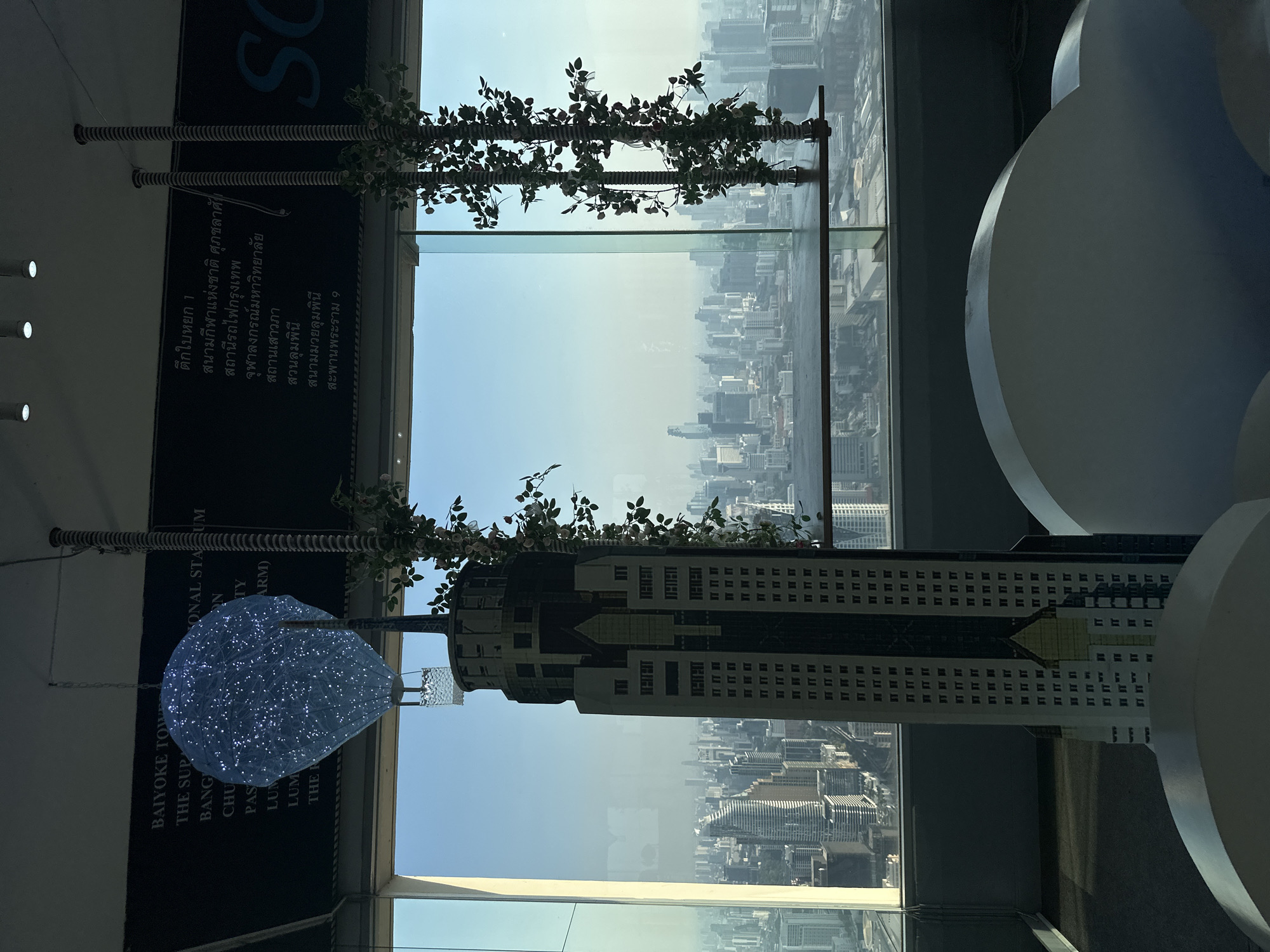 The image shows a view from the Baiyoke Tower in Bangkok, Thailand. The cityscape is visible through a large window, with decorative floral arrangements and a scale model of the building itself in the foreground. A sign with Thai text and English translations indicating locations like "National Stadium," "Bangkok Railway Station," and "Lumphini Park" is visible.