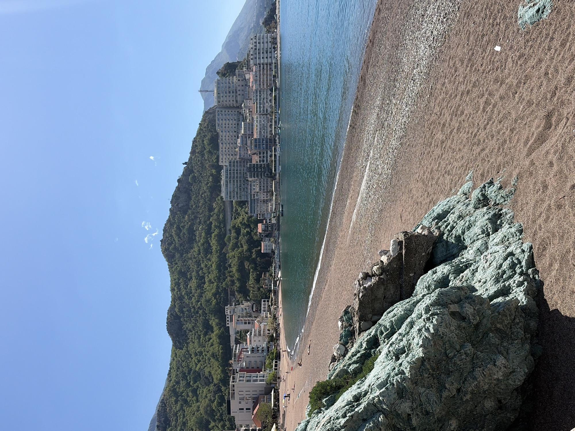 The image shows a coastal scene in Rafailovići, Montenegro, featuring a pebble beach meeting a clear, calm sea. Buildings line the beach and climb up the hillside, indicating a dense urban development. A forested mountain forms the backdrop, adding a natural element to the urban setting. In the foreground, textured, light-green rocks are visible. The sky is a clear, light blue, suggesting a sunny day. The scene includes people walking on the beach and interacting with the sea, and there are boats in the water. The perspective is from an elevated position looking down onto the beach and coastline.