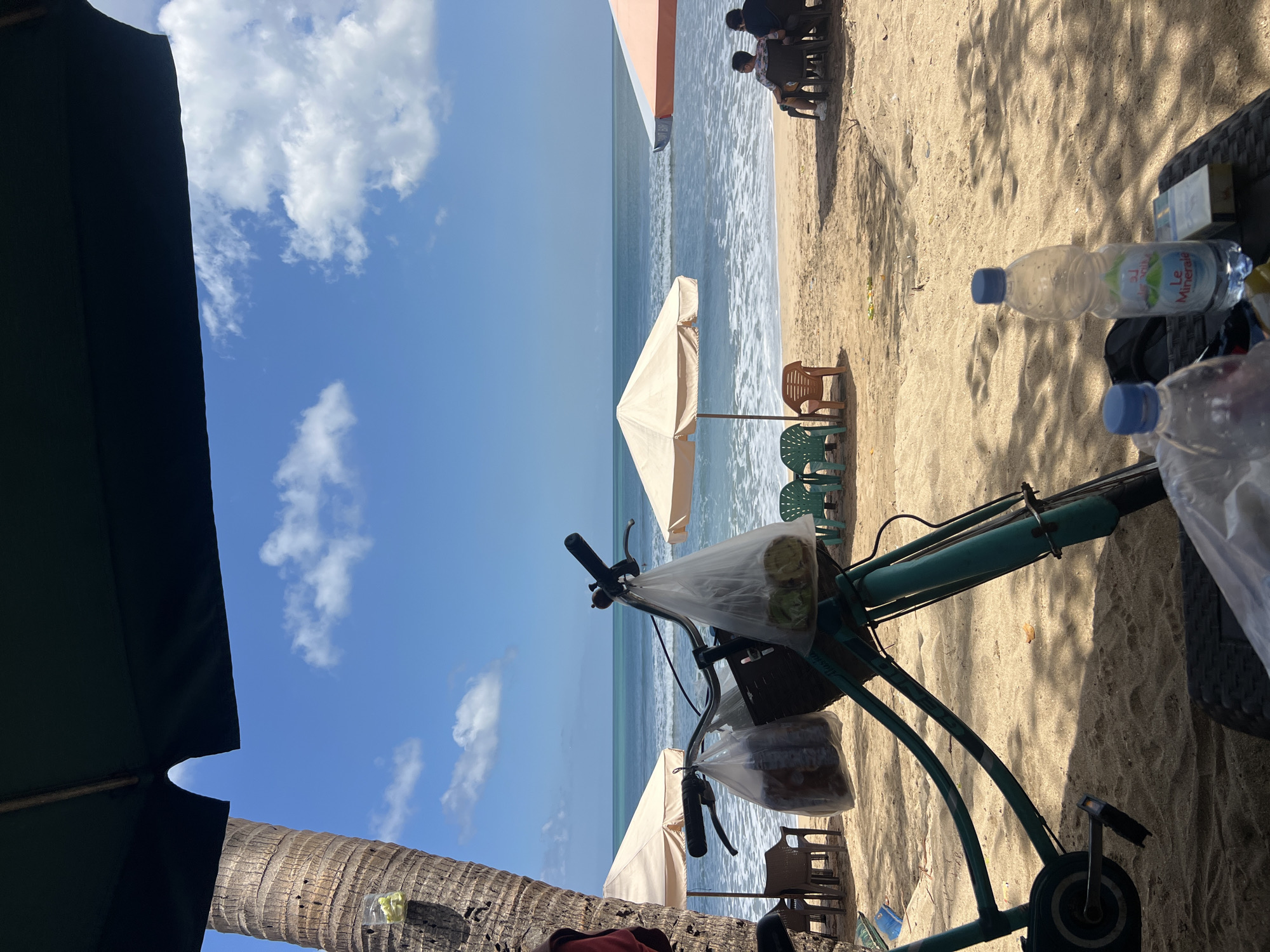 A teal-colored bicycle is parked on a sandy beach in Kuta, Indonesia, with plastic bags containing items hanging from its handlebars. Behind the bicycle, the turquoise ocean meets the horizon under a bright blue sky dotted with white clouds. Beach umbrellas are set up on the sand, with several empty chairs scattered around. Two men are seated under an orange and white umbrella in the background, facing the sea. A palm tree trunk is visible on the left side of the image, and the foreground shows a table with two plastic water bottles and a pack of cigarettes. The scene suggests a relaxed beach atmosphere.