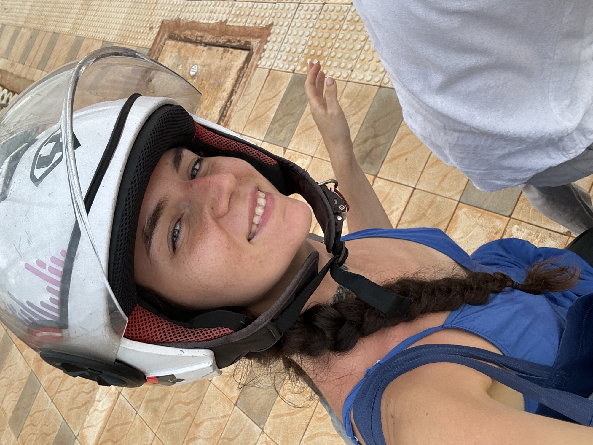 A woman wearing a white helmet and blue dress is standing on a tiled surface. She has dark hair braided over her shoulder and is smiling. A person in a white shirt is beside her, with their arm and torso partially visible. The background features beige tiles. The location is likely Mui Ne, Vietnam, given the style of tiles and the overall aesthetic. The woman appears to be wearing a helmet possibly for riding a motorcycle, and may have stopped to take a selfie or have her picture taken. The tiles indicate an outdoor area, possibly a sidewalk or public space.