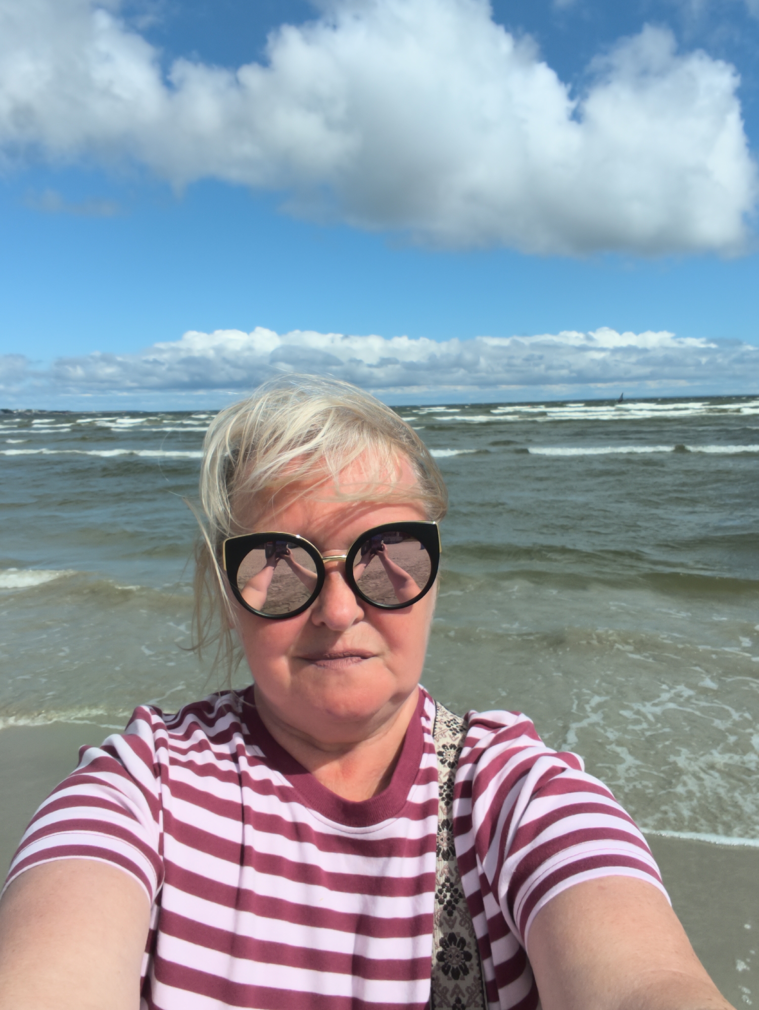 A woman with blonde hair and sunglasses is taking a selfie on a beach. She is wearing a red and white striped t-shirt and a patterned scarf is visible underneath her t-shirt. The ocean is behind her with waves and whitecaps. The sky is blue with white clouds. The location is a beach in Tallinn, Estonia.