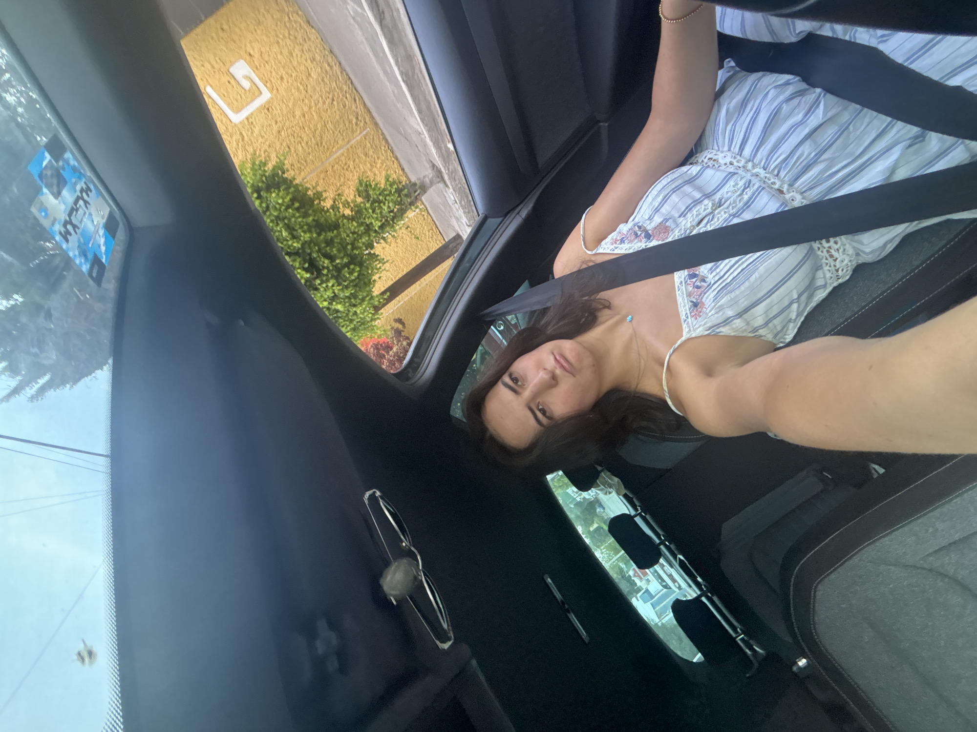 A young woman sits in the passenger seat of a vehicle, buckled in with a seatbelt. She is wearing a striped dress. Outside the car window, there is a yellow building with a white "G" symbol, and a bush. The picture was taken in Puebla, Mexico, based on the license plate sticker visible on the windshield. The picture is taken from the driver's point of view.