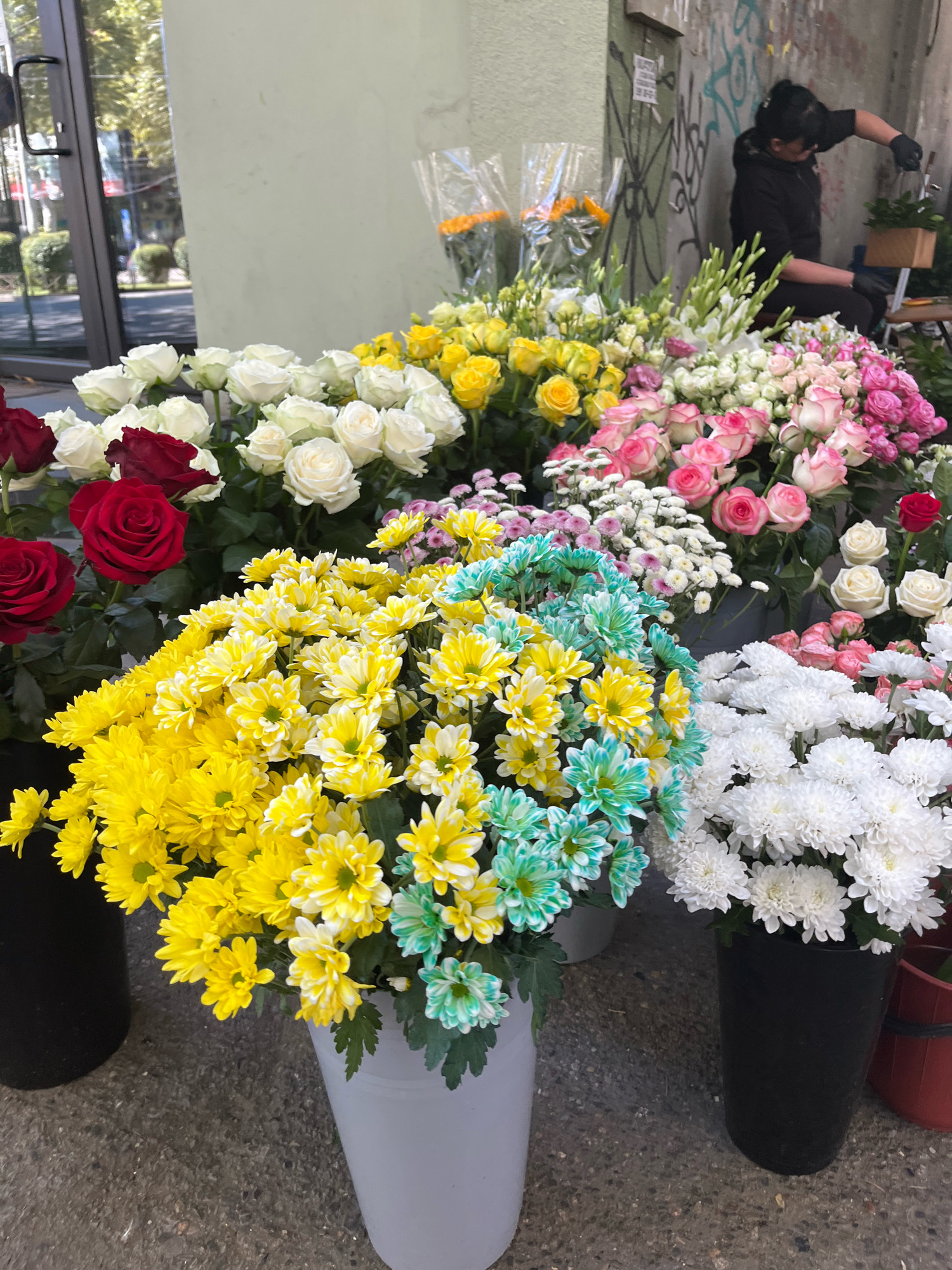 The image depicts a flower stall on a city sidewalk, likely in Tbilisi, Georgia, given the context of similar scenes observed in the city. In the background, a person wearing a black hoodie and gloves is seen working with plants. The foreground is dominated by a vibrant display of various flowers, including red roses, white roses, yellow chrysanthemums, pink and white roses, and a bouquet of turquoise and yellow chrysanthemums. The flowers are arranged in several pots and containers, some of which are black and white. The ground appears to be paved with concrete or asphalt. The overall impression is of a bustling urban environment with a touch of natural beauty.