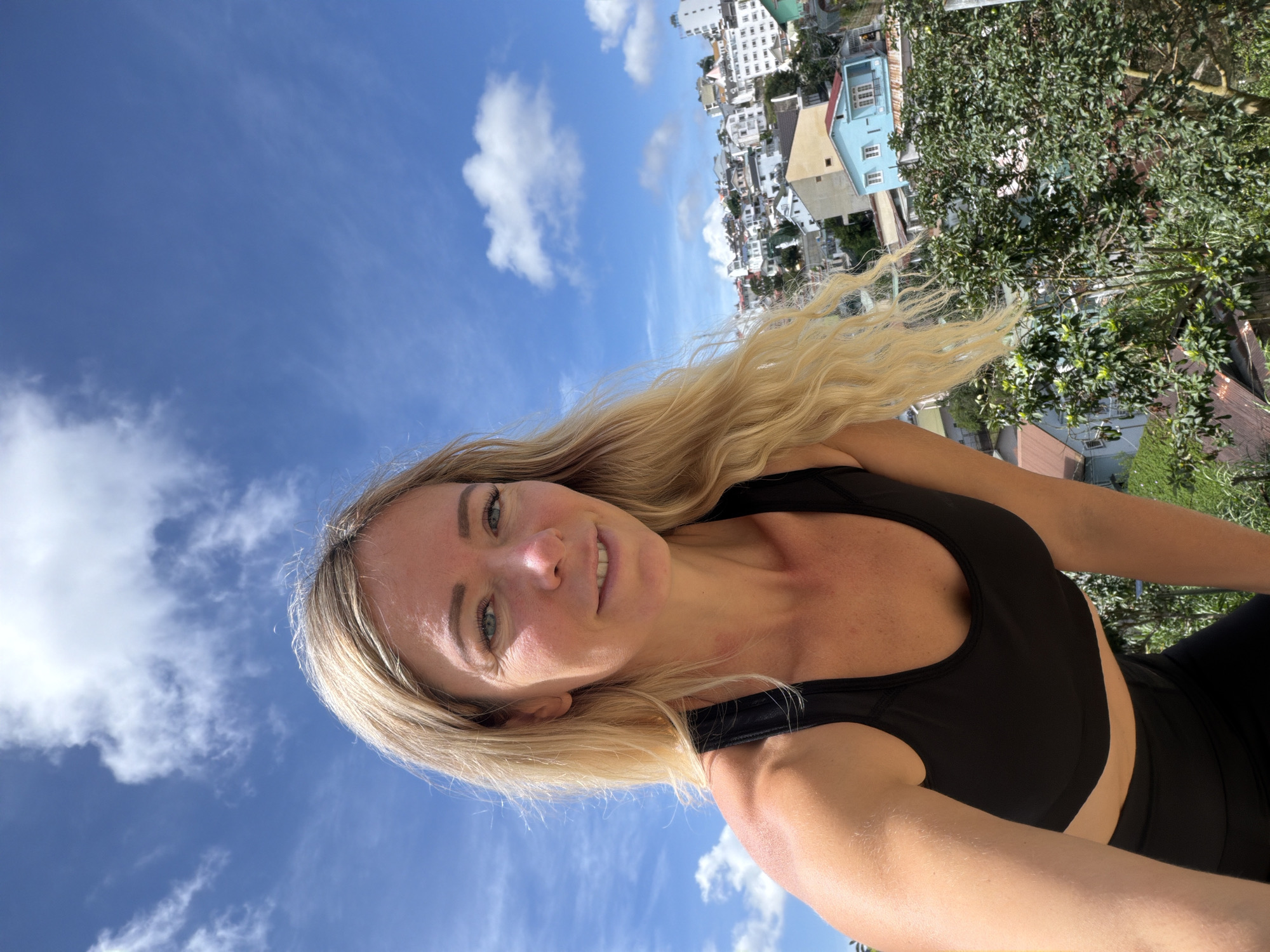 A blonde woman wearing a black sports bra and black leggings poses for a selfie outdoors with a city skyline in the background under a blue sky with white clouds. The woman is leaning forward and smiling at the camera, her blonde hair is flowing. The city appears to be Da Lat, Vietnam, characterized by its hilly terrain and buildings with varying architectural styles and colors. The woman's skin is slightly sun-kissed and she has light blue eyes and fair skin. The overall scene suggests a bright, sunny day.