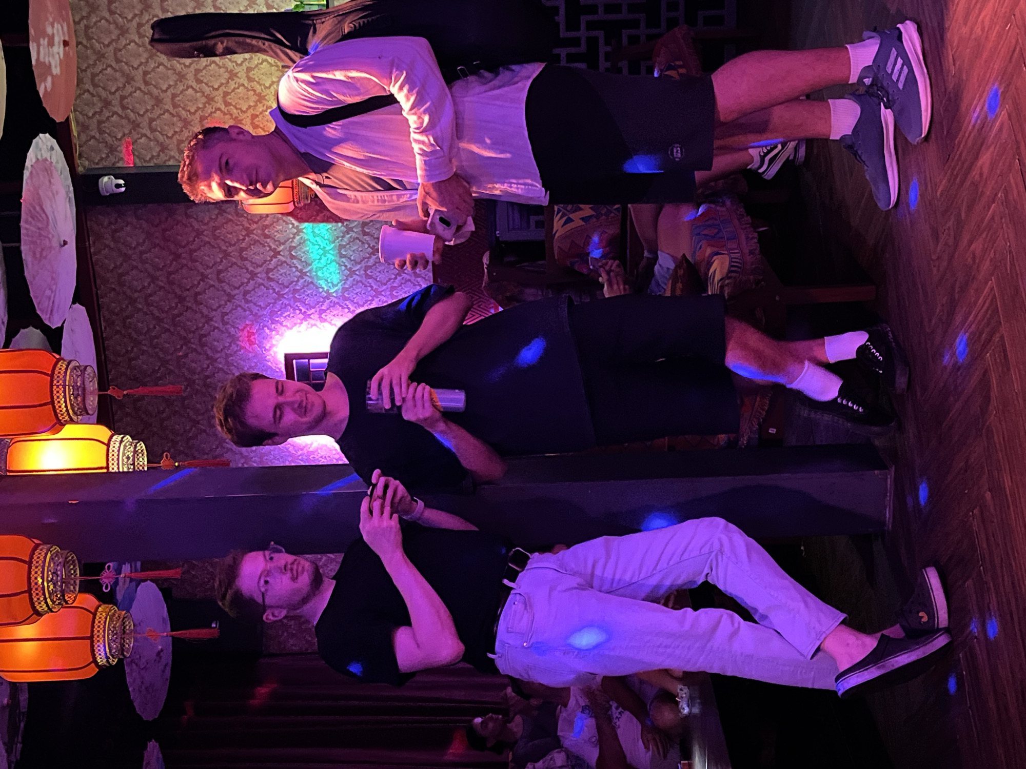 The scene shows a dimly lit bar or lounge in Đà Nẵng, Vietnam, with three young men posing for a photo. The decor includes hanging lanterns, parasols, and wallpaper with an ornate pattern. At the top of the image, a man in a white button-down shirt, black shorts, and gray sneakers has a guitar case slung over his shoulder and is holding a white cup and phone. Below him, a man in a black t-shirt and black shorts holds a silver can and looks at the camera. At the bottom of the image, a man with glasses in a black t-shirt and light-colored pants has a cell phone in his hands. At the bottom of the photo, multiple people sit at tables. The floor is a wooden parquet and is speckled with blue lights.