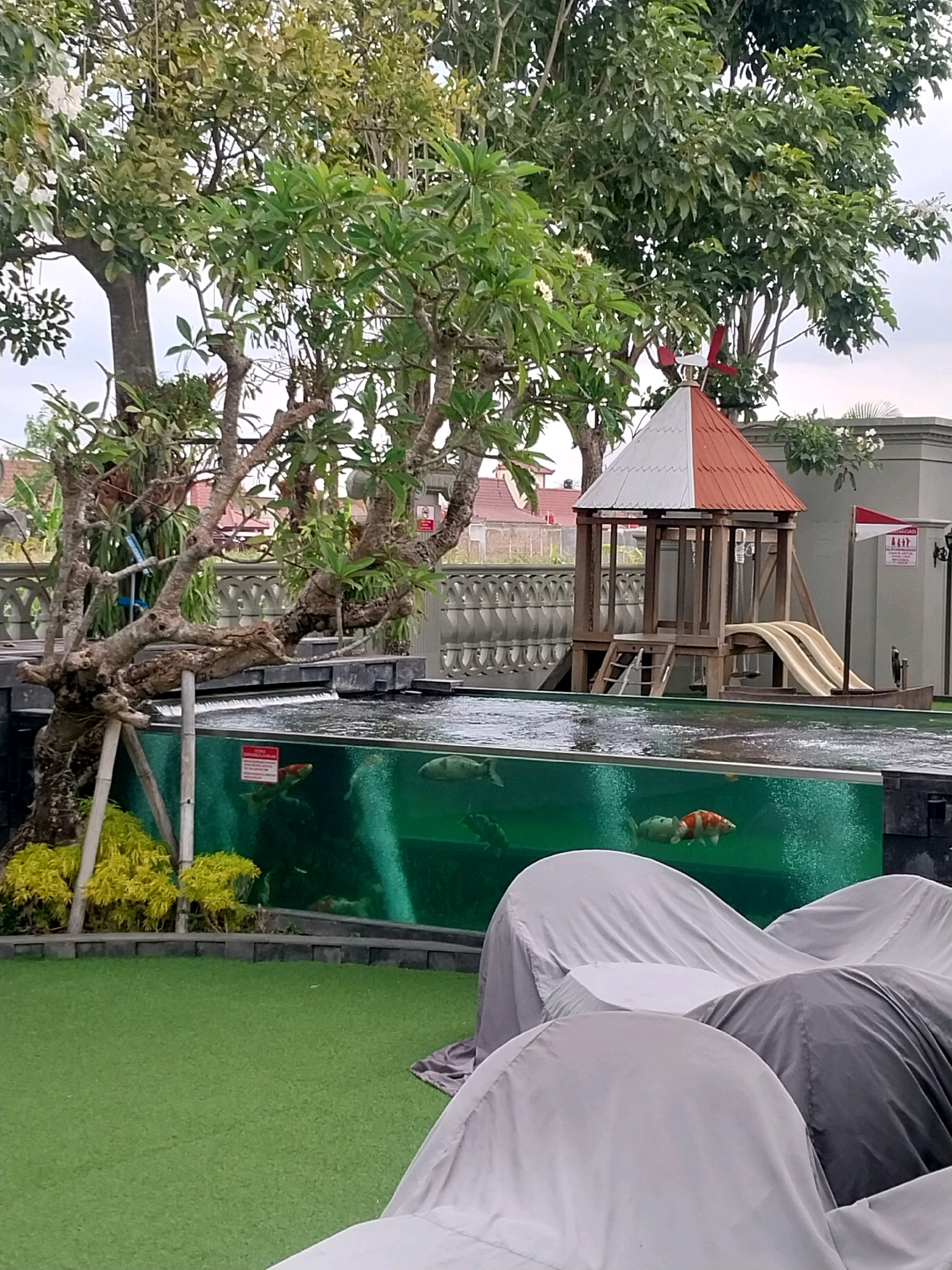 The image shows a residential outdoor area in Bugisan, Indonesia, featuring a koi pond with clear glass walls. Two koi fish, one orange and white and another light green, are visible swimming in the pond. A wooden play structure with two slides and a ladder is situated behind the pond. Lush green trees and foliage surround the area, with a white and red tiled roof structure at the top of the play equipment. The foreground is covered in artificial grass, with several outdoor furniture pieces draped in gray protective covers. The overall impression is that of a well-maintained recreational space, likely part of a private residence or resort.