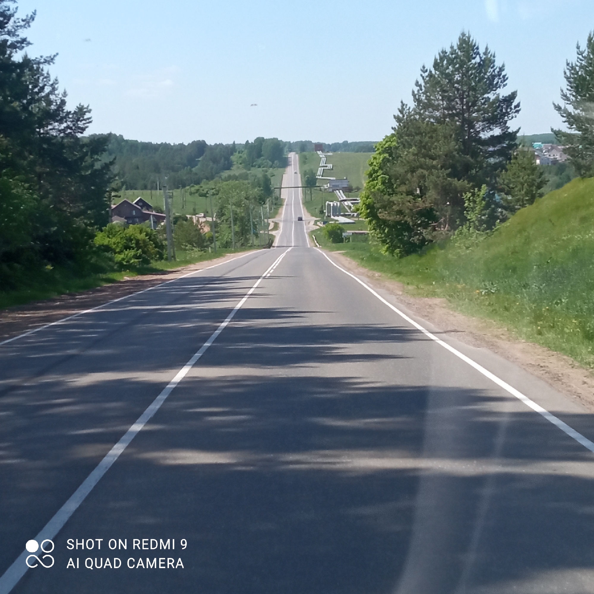 The image captures a straight, two-lane asphalt road extending into the distance, likely a rural or suburban setting near Прикамский, Russia. White lines delineate the lanes. The road ascends a slight hill, obscuring the view beyond. Shadows cast by trees obscure the asphalt surface. Vegetation lines the sides of the road, including trees and grassy slopes. Powerlines run alongside the road. In the distance, pipelines can be seen above ground. A few buildings are visible on either side of the road, suggesting scattered dwellings. A vehicle is visible on the road in the distance. The sky is clear and blue. The weather appears to be sunny.