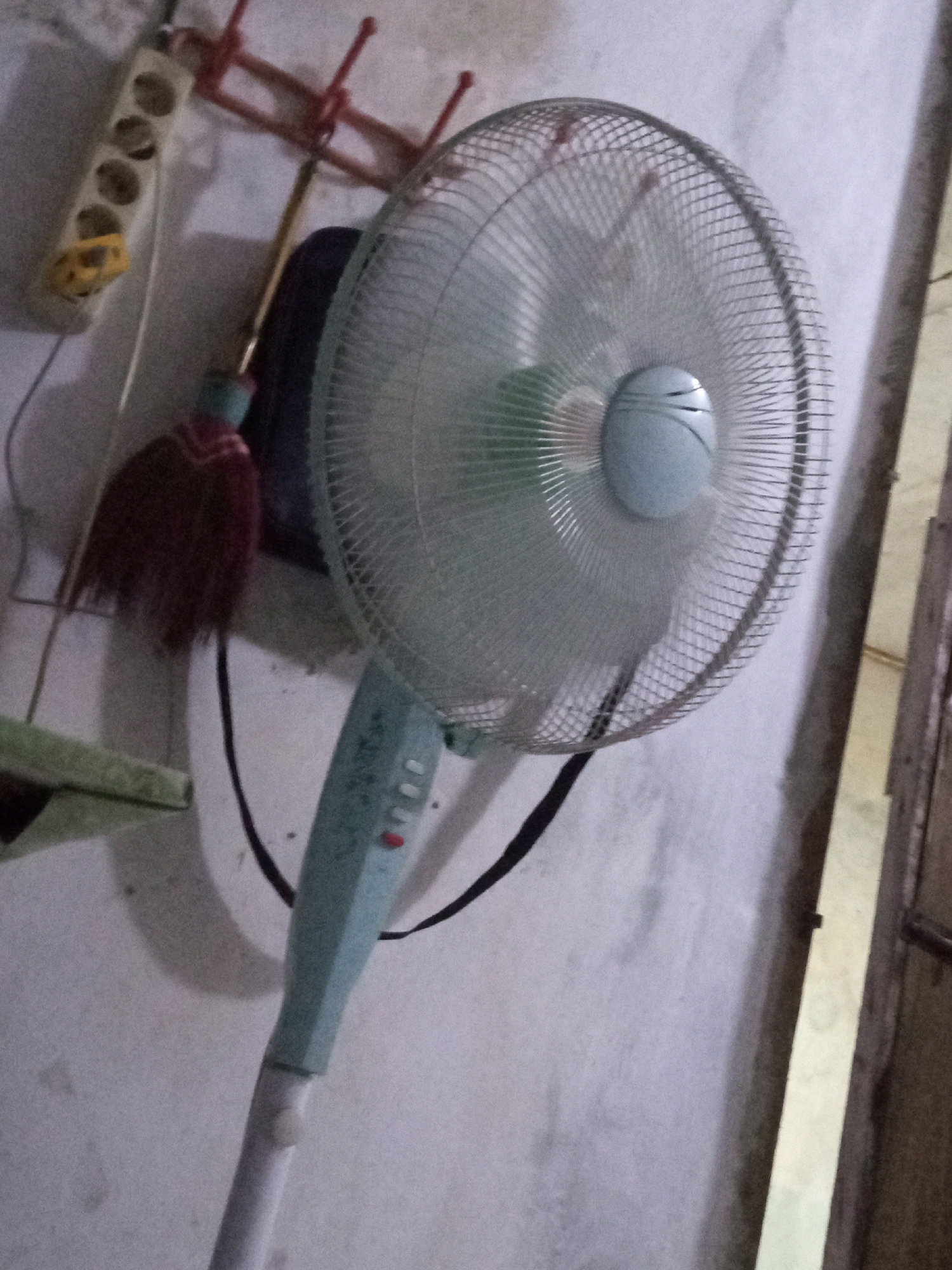 The image shows a fan in a room. The fan is light blue and has a mesh grill. It is on a stand and appears to be plugged into a power strip mounted on the wall. There are some items hanging on the wall behind the fan, including what looks like a broom and a red rack with hooks. The wall is a light color and appears to be textured. The lighting in the room is dim, suggesting it may be nighttime or the room is not well-lit. There is no visible indication of the city of Boyoteluk, Indonesia in the image.