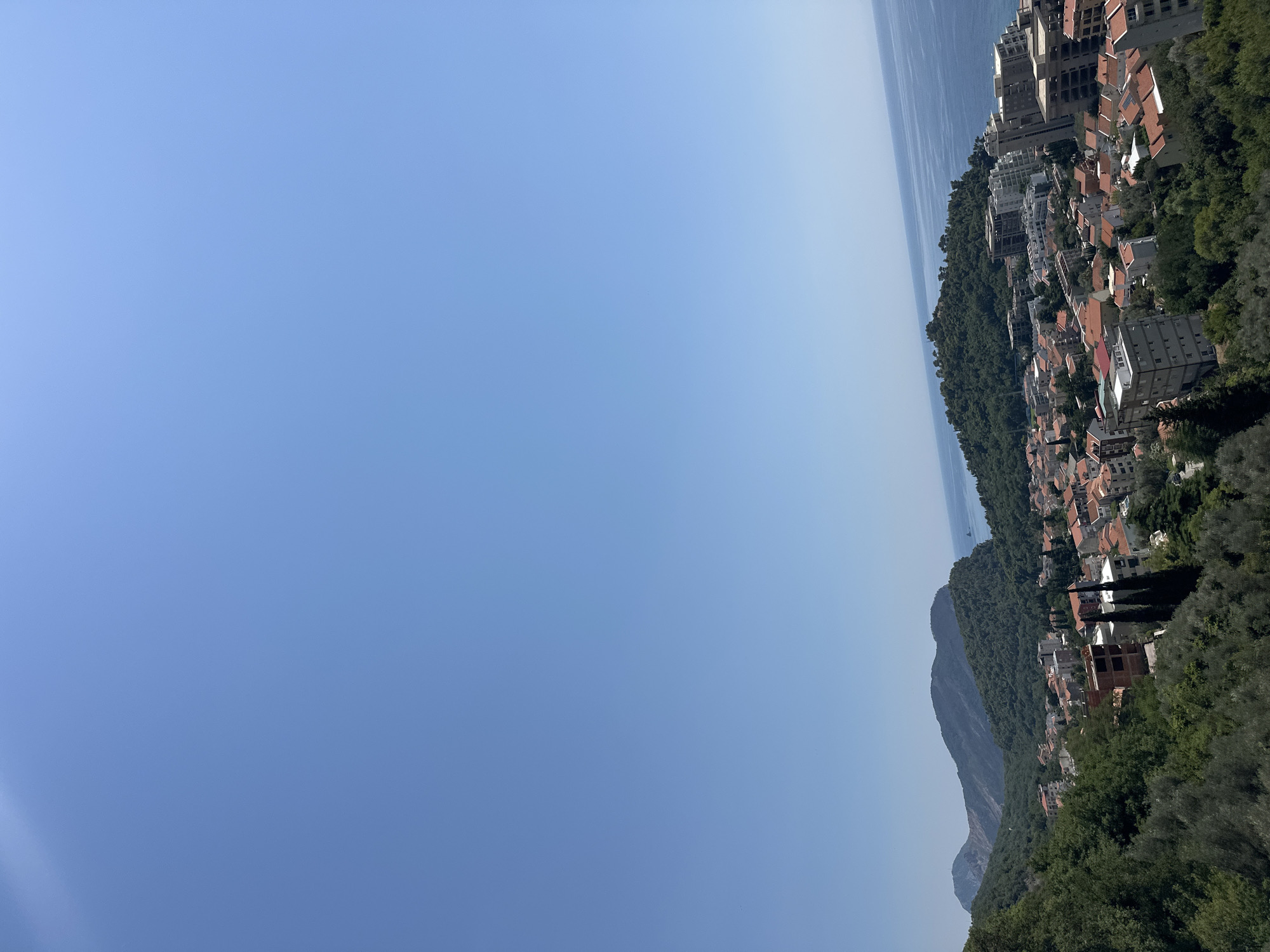 The image depicts a panoramic view of the city of Stupovi, Montenegro, under a clear blue sky. In the foreground, lush green trees and foliage blanket a hillside. Below, a dense cluster of buildings with terracotta roofs spreads across the landscape. These structures range from multi-story apartment buildings, some appearing modern and unfinished, to smaller residential homes. The city extends towards the coast, where the deep blue Adriatic Sea meets the horizon. In the distance, rolling, tree-covered mountains frame the scene. The image captures a sunny, tranquil day in a coastal Montenegrin town.