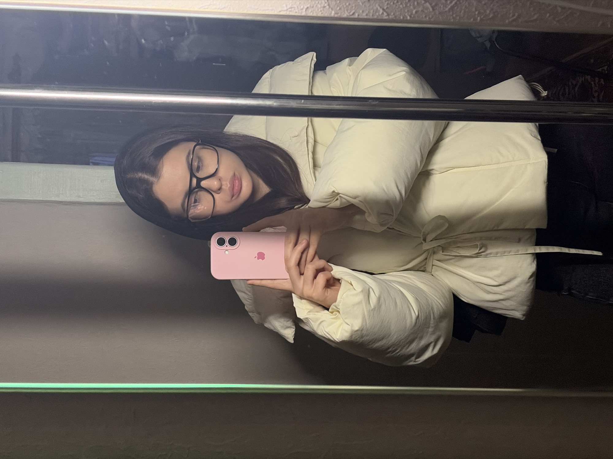 The media file displays a mirror selfie of a young woman, rotated 90 degrees clockwise. The woman is positioned horizontally across the frame, appearing to lie on her back and look upwards. She has long dark hair and wears black-rimmed glasses. Her lips are slightly parted. She is dressed in a light-colored, possibly cream or pale yellow, puffy jacket or coat.
In her hands, she holds a pink smartphone, identified as an iPhone by the visible Apple logo on its case, pointing it towards the mirror she is captured in. Her gaze is directed towards the phone's screen or her reflection within it.
A prominent, reflective metal bar runs horizontally across the middle of the image. Above this bar, the mirror reflects a dark and somewhat indistinct background, possibly indicating a cluttered area or a room with low lighting. Below the bar, the background appears to be a plain wall, with a faint green-hued line near the bottom edge, which could be part of a fixture or reflection.
The overall lighting is subdued, with brighter reflections concentrated on the metal bar. The activity is clearly a mirror selfie being taken indoors in Chaikovsky, Russia. There is no other discernible text apart from the Apple logo on the phone.