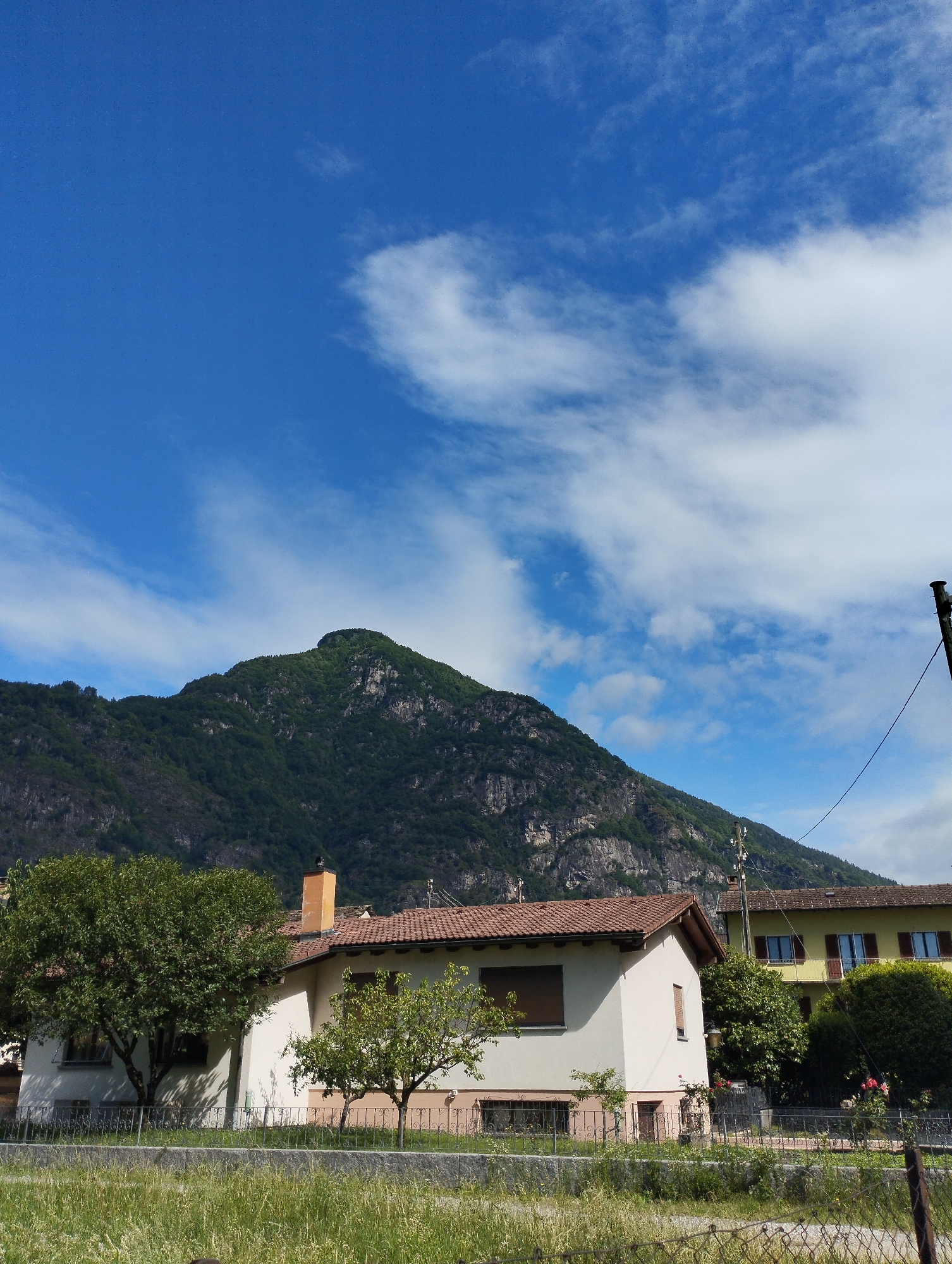 In this sunlit outdoor scene, a sprawling, verdant mountain dominates the background under a bright blue sky scattered with wispy white clouds. In the middle ground, a cluster of buildings sits at the base of the mountain. A white house with a red-tiled roof and a prominent orange chimney is situated to the left. To its right, a yellow house with a similar red-tiled roof is visible. Both houses appear to be residential, with windows and shutters. In the foreground, a chain-link fence borders a grassy area, with lush green grass and some wildflowers. Trees, including a large deciduous tree to the left of the white house and a smaller fruit tree in front of it, are present. A power pole with visible wires stands to the right of the yellow house. The scene appears to be a peaceful rural or suburban setting.