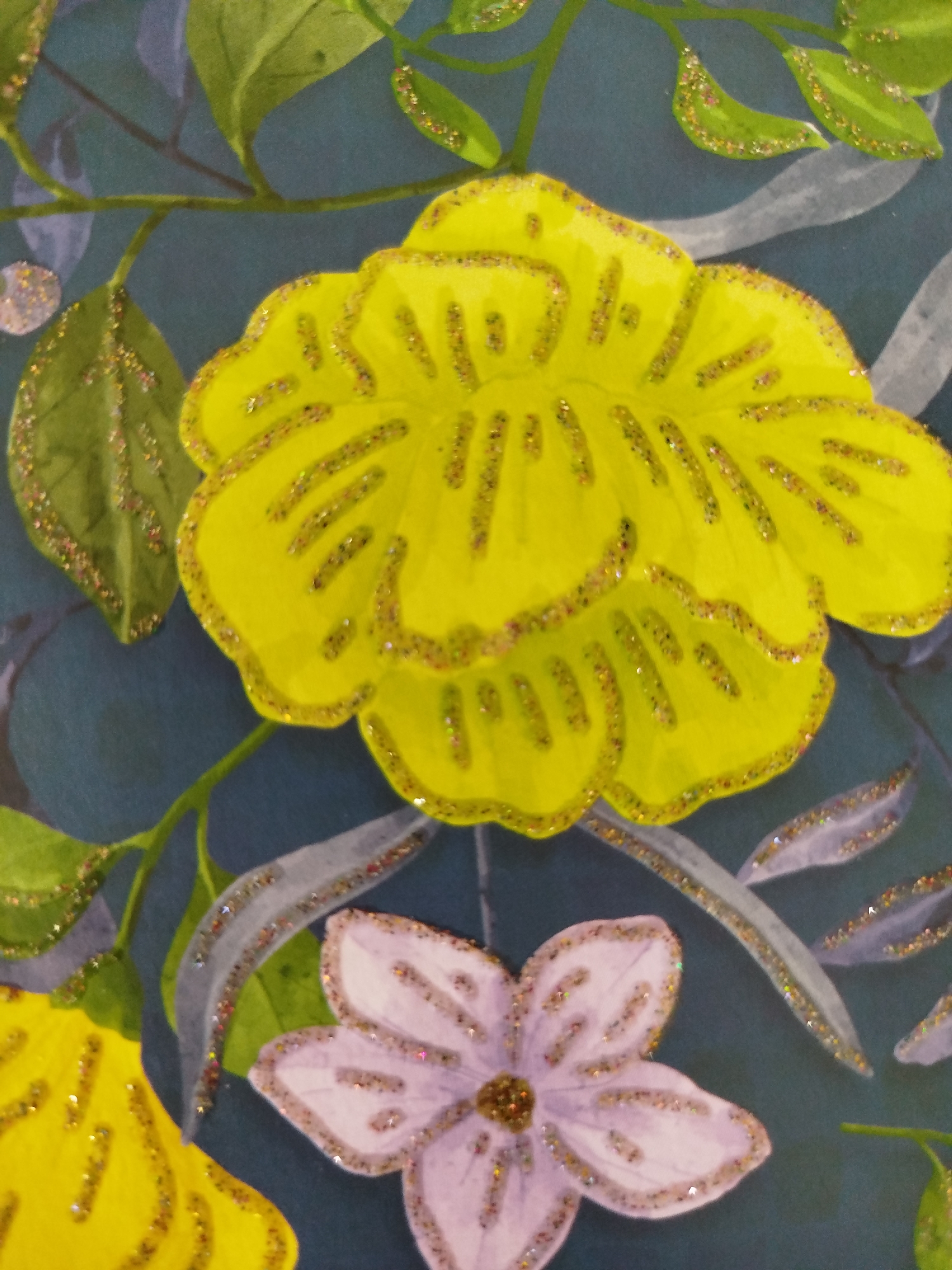 The image is a close-up of a painting featuring stylized flowers and leaves. The background is a dark, muted teal. A large, vibrant yellow flower with scalloped edges dominates the upper portion of the frame, outlined with glitter. To the left, several green leaves are visible, also with glitter accents. Below the yellow flower, a smaller, pale lavender flower with five petals is depicted, also outlined in glitter. Long, wispy grey shapes, possibly abstract foliage or branches, are scattered throughout the background, some with glitter. A thin green stem or branch extends from the upper left corner, weaving through the composition. The overall impression is one of a decorative, possibly handmade artwork. There is no discernible location in the city of Крывасельскі сельскі Савет, Belarus, as this is a still image of a painting.