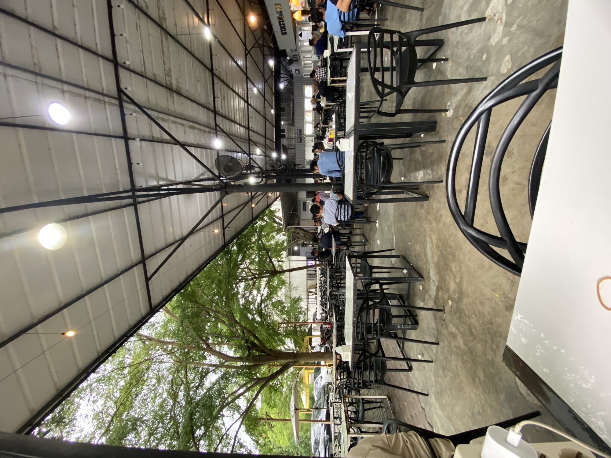 The image captures a lively, semi-outdoor restaurant or food court setting in Lhokseumawe, Indonesia, likely during the evening or dusk. The scene is dominated by a vast, high metal roof structure supported by black beams and pillars, from which numerous bright lights and decorative string lights illuminate the area. Several industrial fans are also mounted on the pillars.
In the foreground, the edge of a white table is visible on the right, with a modern black chair beside it. A white power adapter with a cable rests on the table, suggesting charging a device. The floor is a textured concrete surface.
The midground features numerous white-topped tables and black chairs, many of which are occupied by patrons. Several people are seated in small groups, appearing to be dining, talking, or waiting for food. Their attire is casual. Some chairs are stacked, perhaps indicating additional seating or areas being prepared.
To the left, a prominent feature is a large, mature tree with abundant green foliage, its branches extending over part of the dining area and reaching towards the roof line, adding a natural element to the otherwise industrial setting. Beyond the tree, glimpses of parked cars and low-rise buildings are visible.
In the background, under the far end of the roof, more people are seated at tables. A sign is clearly visible that reads "STATION" with "FOOD" partially visible below it, likely indicating the name of the establishment or a specific food stall. The overall atmosphere appears casual and bustling.