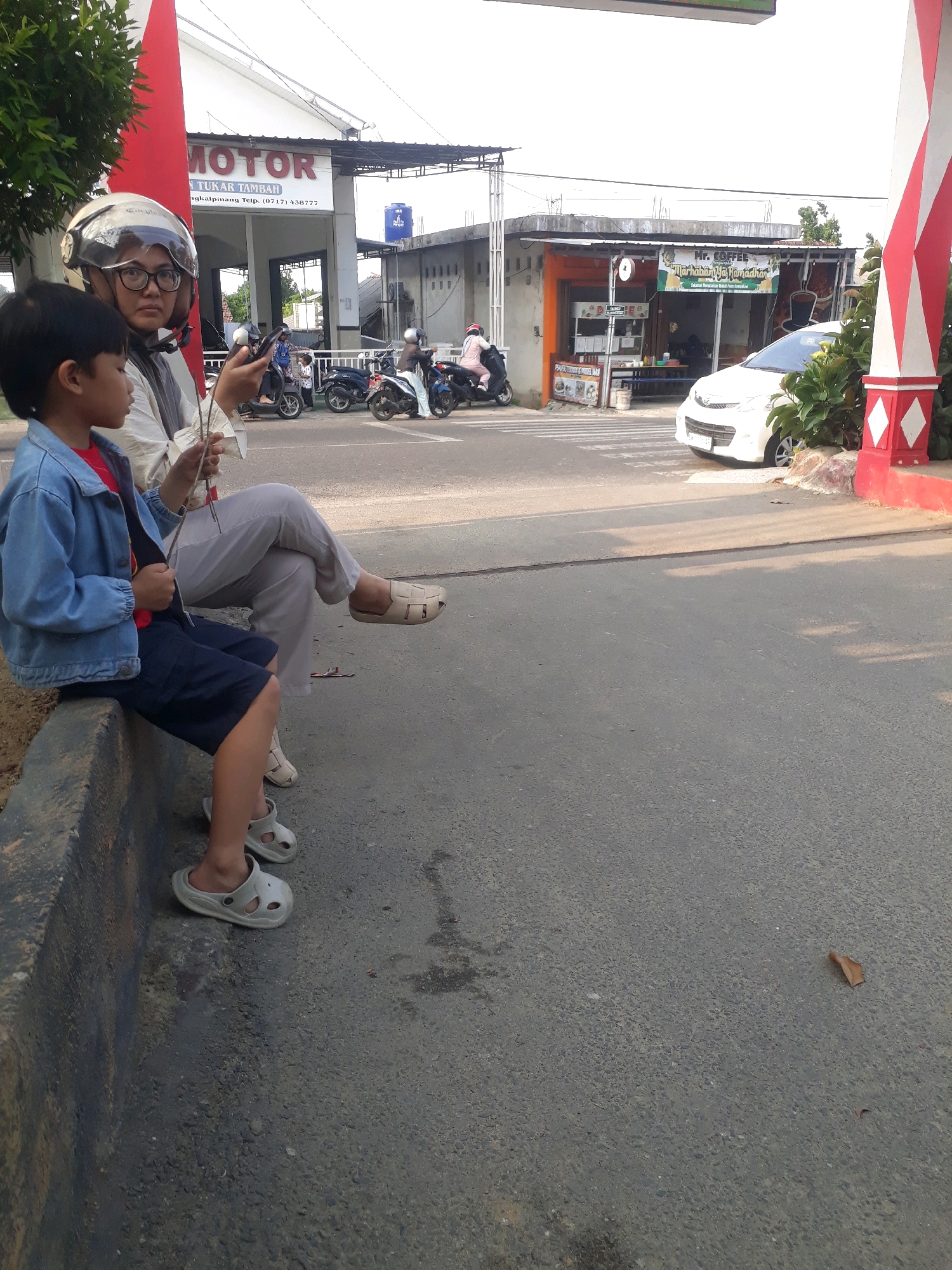 A woman wearing a helmet and glasses sits on a concrete curb next to a boy. The woman wears a light-colored top and gray pants. The boy wears a blue denim jacket and dark shorts. He has white sandals on his feet. The woman holds a phone and a piece of paper with sticks in her hands. Behind them, several motorbikes are parked. In the background, there is a building with a sign that reads "MOTOR" in red letters. To the right, there is another building with a sign that says "Mr. COFFEE". A white car is parked in front of the coffee shop. The scene appears to be a street in Pangkalpinang, Indonesia, with the presence of local businesses and transportation common to the region.