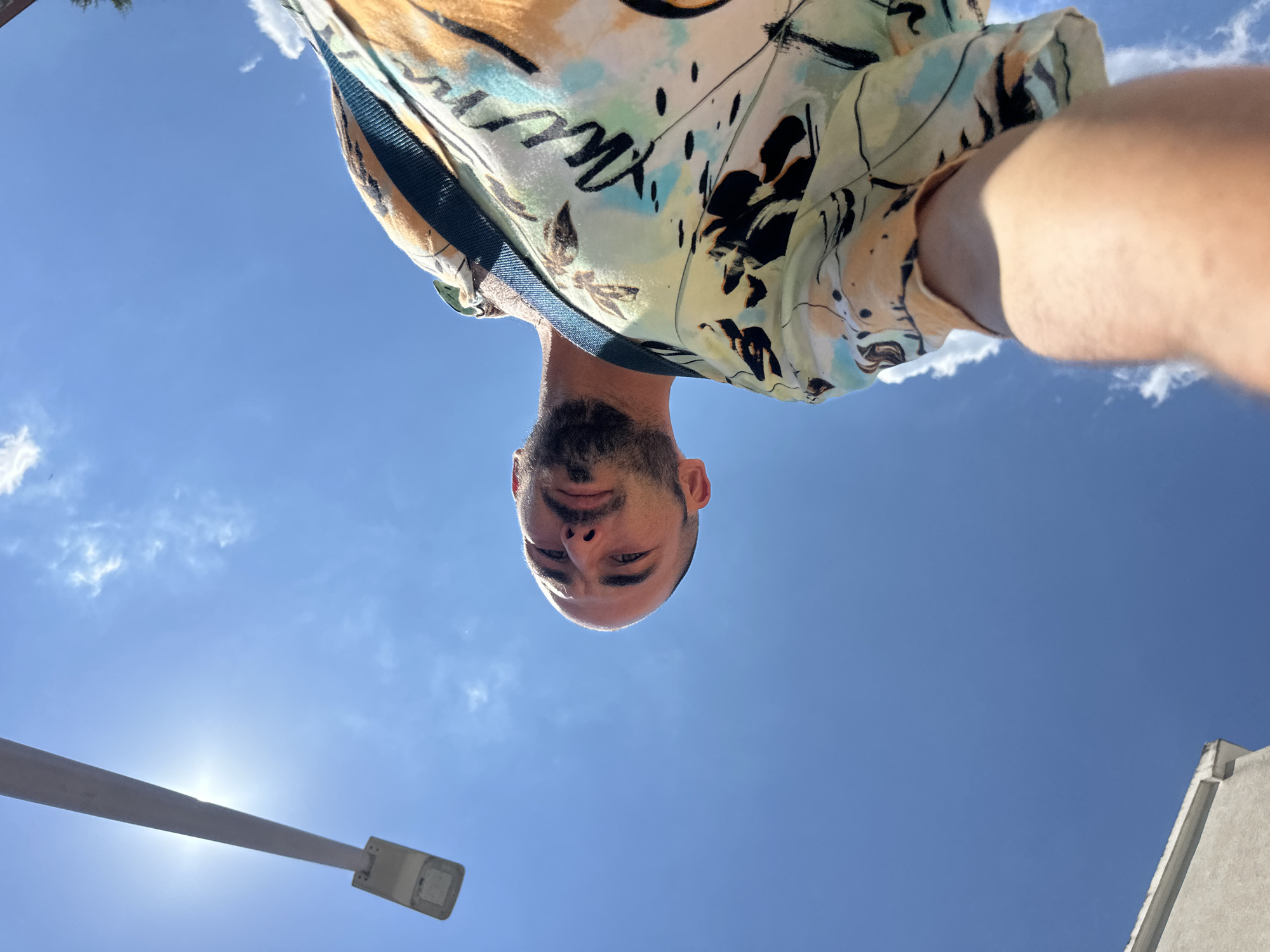 A low-angle, wide shot captures a man posing for a selfie against a bright blue sky with scattered clouds, on a sunny day in Budva, Montenegro. The man, facing the camera from a slightly upward perspective, has a shaved head and a beard. He is wearing a vibrantly patterned short-sleeved shirt in shades of yellow, orange, turquoise, and black, with a dark strap across his shoulder. In the upper left of the frame, a street lamp extends diagonally across the sky, its light source glowing intensely. A portion of a building with a white cornice is visible in the lower left corner, suggesting an urban environment. The overall impression is of a casual, outdoor setting, possibly a street or public area within Budva.