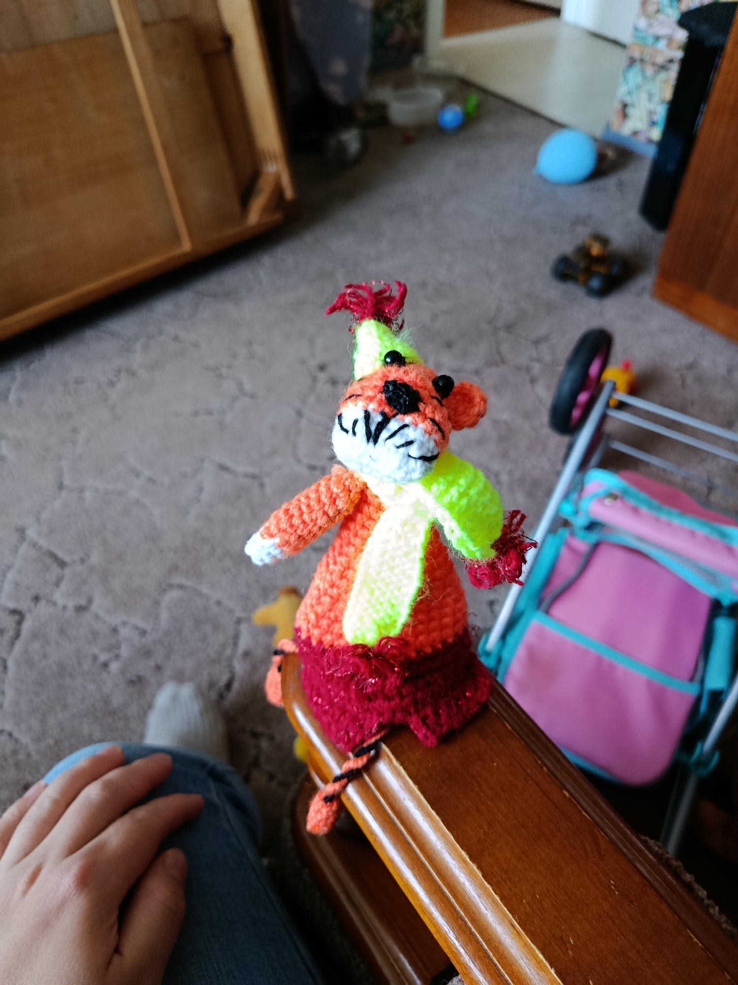 A knitted toy animal with orange and green accents sits on a wooden surface. The toy has black eyes and a striped face. A pink and turquoise bag is visible in the background, attached to a stroller. A person's hand and knee are in the foreground, wearing blue jeans and a grey sock. The floor is carpeted. The scene is indoors, and based on the context of the city of Apatity, Russia, this is likely a domestic indoor setting.