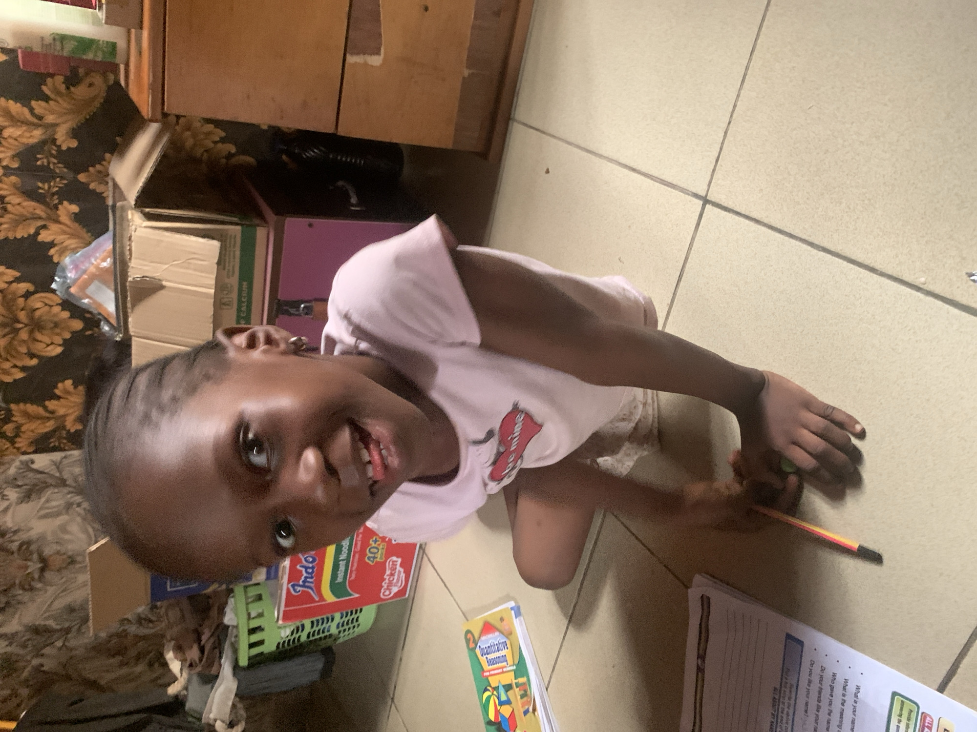 A young Black girl with braided hair and hoop earrings is sitting on a tiled floor, looking directly at the camera with a playful expression, sticking her tongue out slightly. She is wearing a light pink t-shirt with a red heart graphic. In front of her, a workbook titled "All About Names" is open, and she is holding a pencil with her right hand. The background shows a cluttered domestic scene, including a green basket with various items, a red box of "Indomie" instant noodles, cardboard boxes, and furniture, suggesting a home environment in Sabon Tasha, Nigeria. The lighting is warm and appears to be natural light from an unseen source.