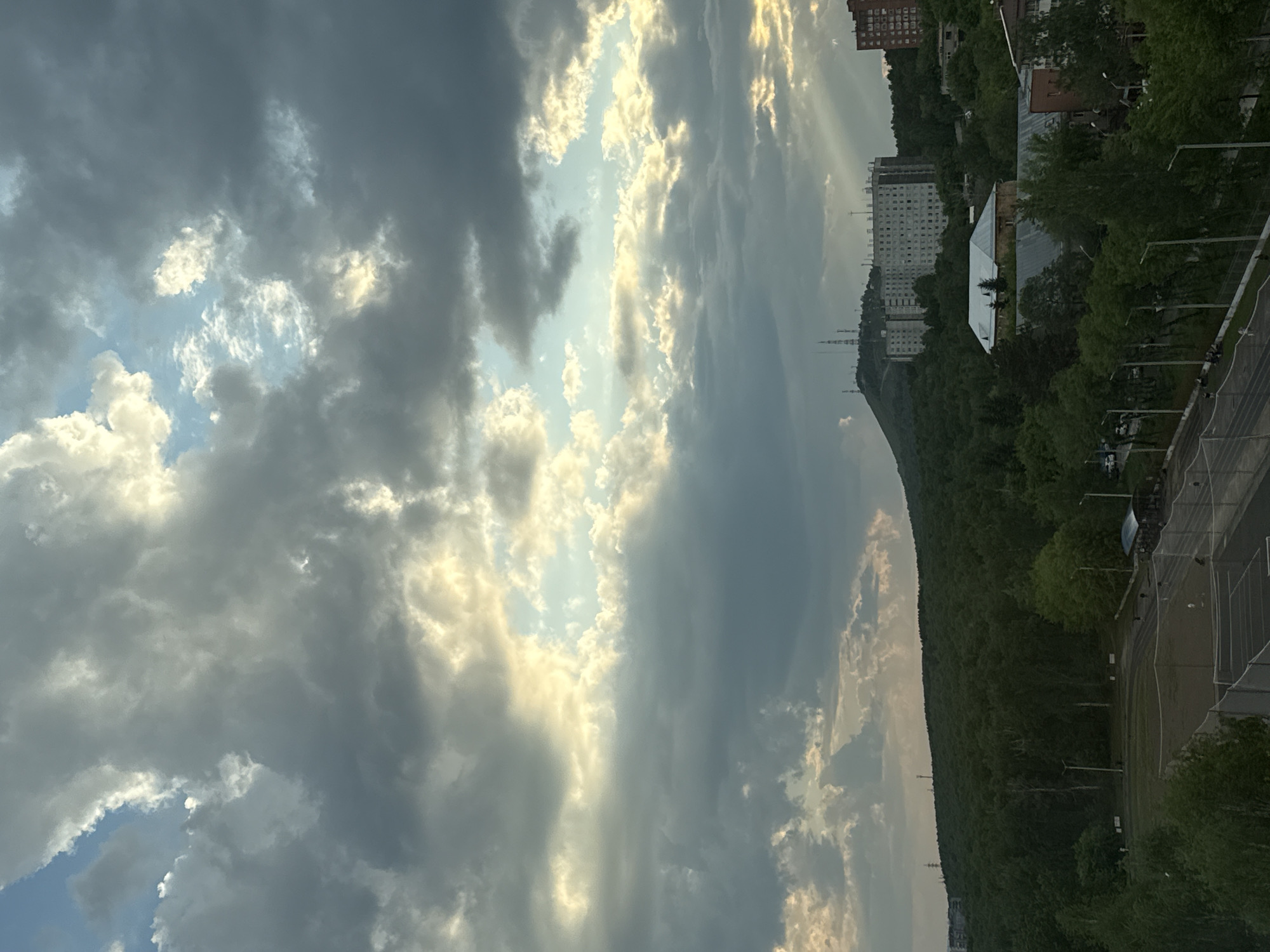The image depicts a sky filled with dramatic clouds, interspersed with patches of bright sunlight. These clouds appear to be a mix of dark, heavy formations and lighter, wispy ones. The sunlight creates a luminous effect, highlighting the textures and shapes of the clouds.
Below the sky, a landscape unfolds, characterized by a densely forested hill sloping downwards. The trees are lush and green, suggesting a temperate season, likely summer or late spring. Along the lower part of the image, a hint of urban development is visible. Several multi-story buildings, typical of Soviet-era architecture, are situated on or near the hillside. One prominent building is a large, light-colored block with many windows. Further down, a white, tent-like structure is visible, possibly a temporary venue or market.
The scene also includes elements of infrastructure and recreation. A road or pathway runs alongside the forested hill, and beyond that, a sports field or stadium with fencing is apparent. Streetlights are also visible, suggesting the presence of an urban or semi-urban area.
The lighting suggests that it is either early morning or late afternoon, with the sun low on the horizon, casting a warm glow through the clouds. The overall mood is somewhat dramatic due to the strong contrast between the dark clouds and the bright sunlight, but also peaceful, given the natural landscape. There is no discernible text in the image. The setting appears to be a natural area bordering a city, likely on a hill overlooking the urban sprawl.