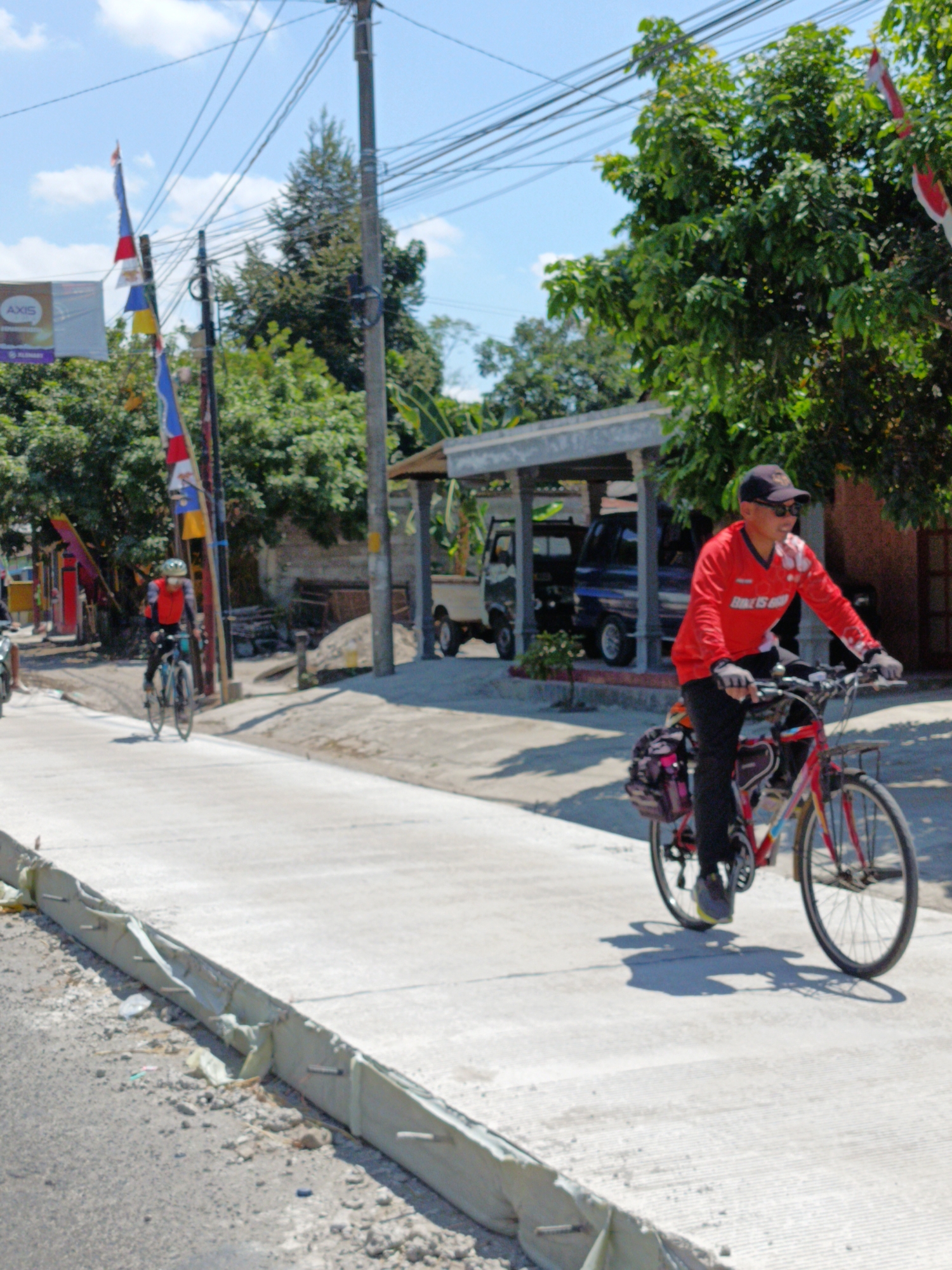 The image depicts a bright, sunny day in Manisrenggo, Indonesia, with a clear blue sky and scattered clouds. The scene is an outdoor street environment, likely a road or pathway, with a paved surface on the right and a rougher, unpaved shoulder on the left.
The primary subjects are two individuals cycling. In the foreground, a man wearing a red long-sleeved shirt, black pants, a black baseball cap, and sunglasses is riding a red bicycle. The bicycle has a rack on the back with bags attached. He is smiling and looking forward. Behind him and to the left, another cyclist, wearing a red vest over a long-sleeved shirt and a helmet, is also riding a bicycle. This cyclist is further away and less detailed.
The background features lush green trees and vegetation. Several utility poles with numerous wires crisscross the upper portion of the image. Decorative flags, primarily red and white and blue and yellow, are visible on poles near the left side of the frame, suggesting a festive or national occasion. A building with a covered structure, possibly a carport or small shop, is present in the middle ground, with a light-colored truck parked underneath. Construction materials, including sand and possibly wood, are visible near the building. A billboard with the word "AXIS" is also discernible in the distance on the left. The overall impression is of a typical day in a somewhat rural or suburban area of Indonesia, with everyday activities like cycling taking place alongside local infrastructure and natural elements.