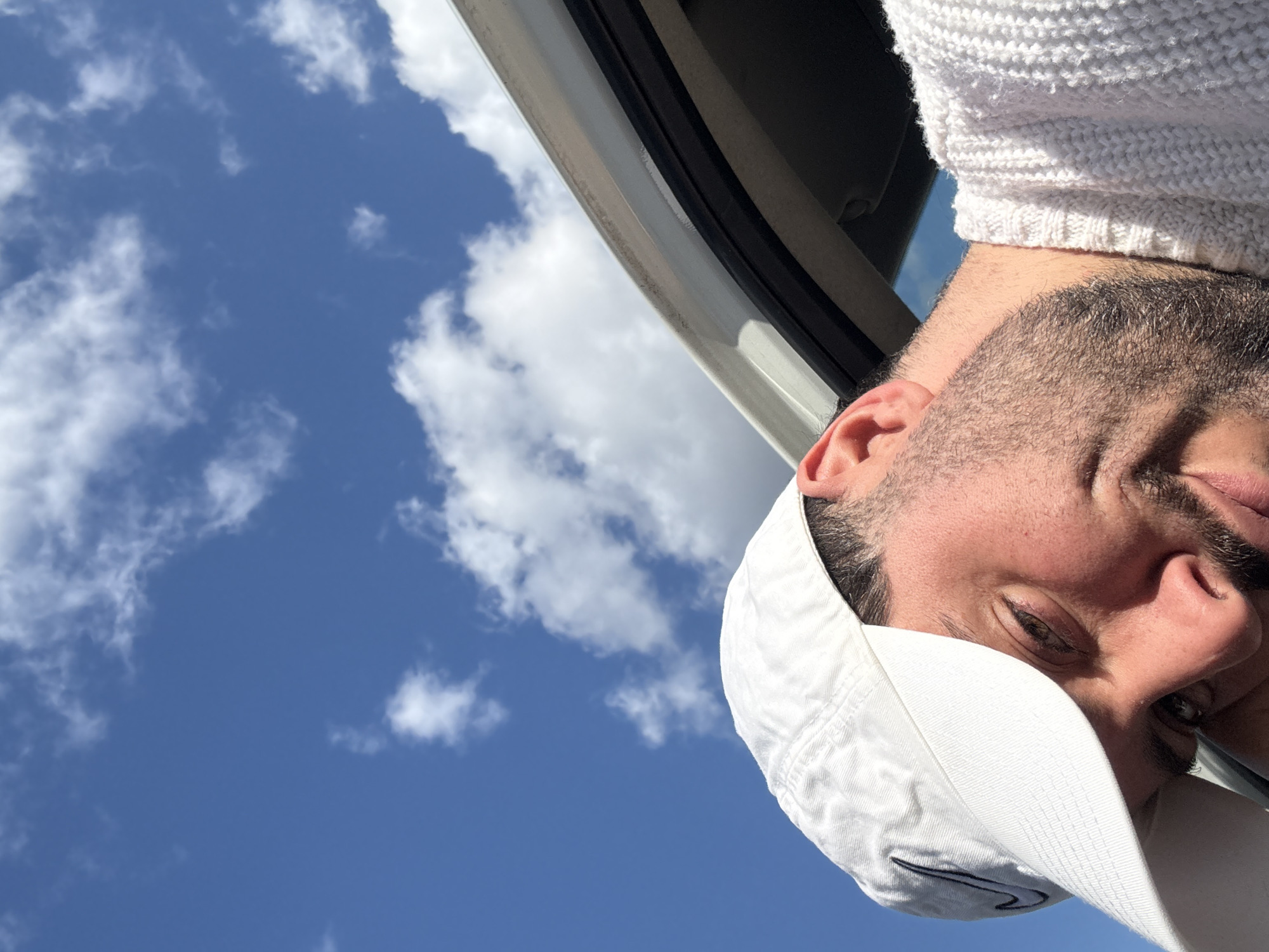 A selfie of a man is taken in Esch-sur-Alzette, Luxembourg. The man wears a white baseball cap with a black Nike logo and a white knit sweater. He has a beard. A car's white doorframe is partially visible in the upper left, with a blue sky and scattered clouds in the background.