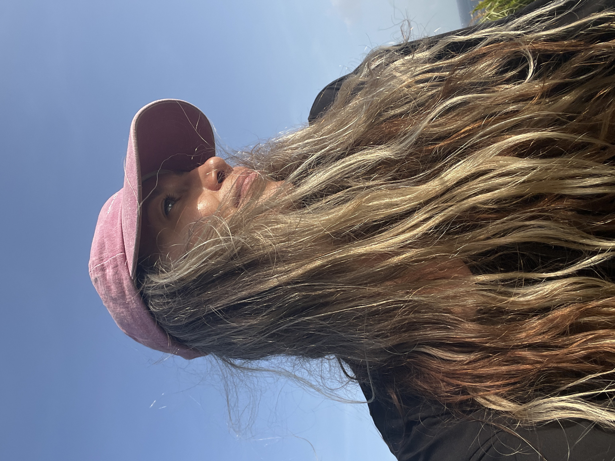 In Dondra, Sri Lanka, a woman with long, wavy, sun-kissed hair and a pink baseball cap is captured in a close-up shot. She is wearing a black top and is looking to her right, with her face partially illuminated by sunlight. The background is a clear blue sky, suggesting an outdoor setting.